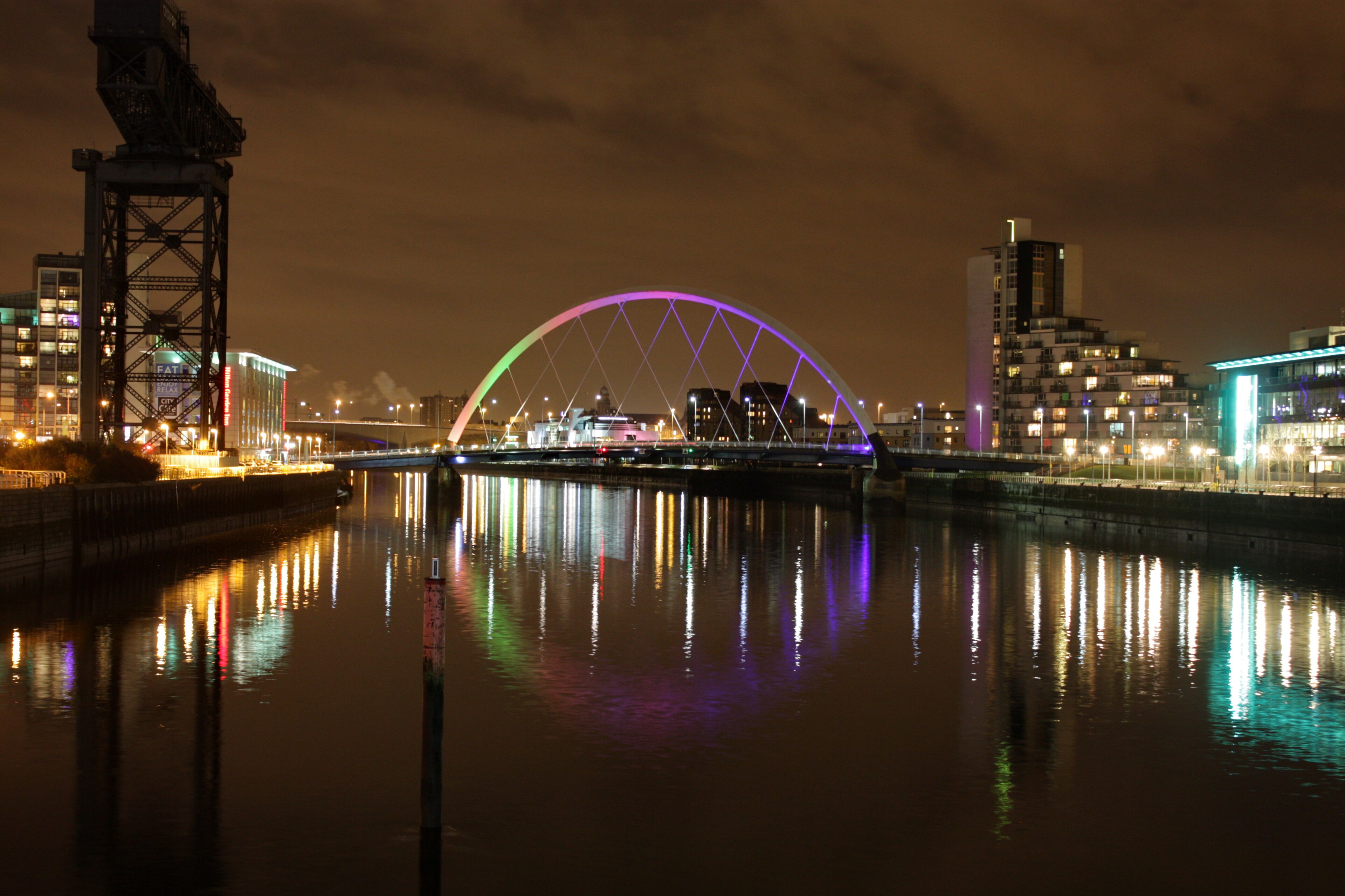 A cityscape at night in Glasgow featuring a brightly lit arch bridge with colorful lights reflecting on the river below, surrounded by modern buildings, illuminated waterfront areas, and plenty of things to do nearby.