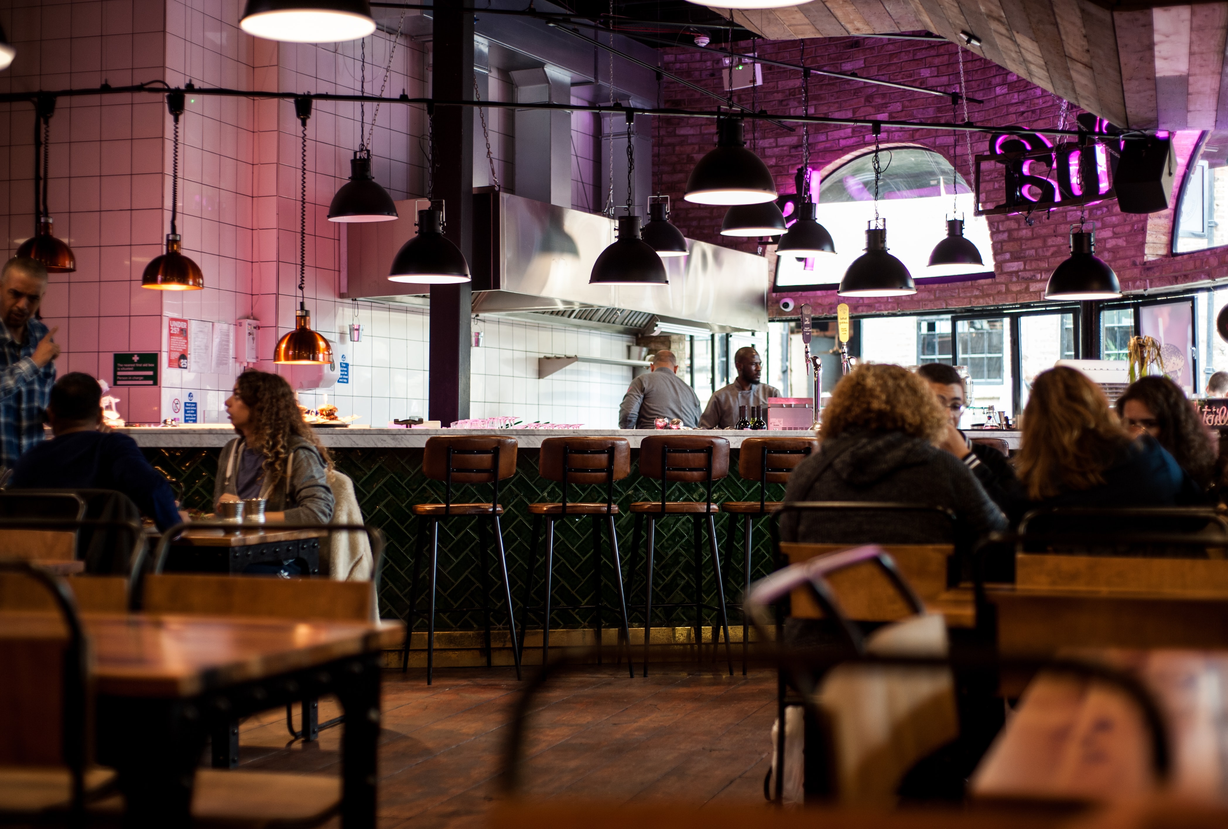 A cozy, modern café located in one of the hottest food spots—Vita Student Lewisham. With wooden furniture, hanging pendant lights, and a brick wall, the warm and inviting atmosphere is complemented by two chefs working in an open kitchen as people sit at tables or the bar.
