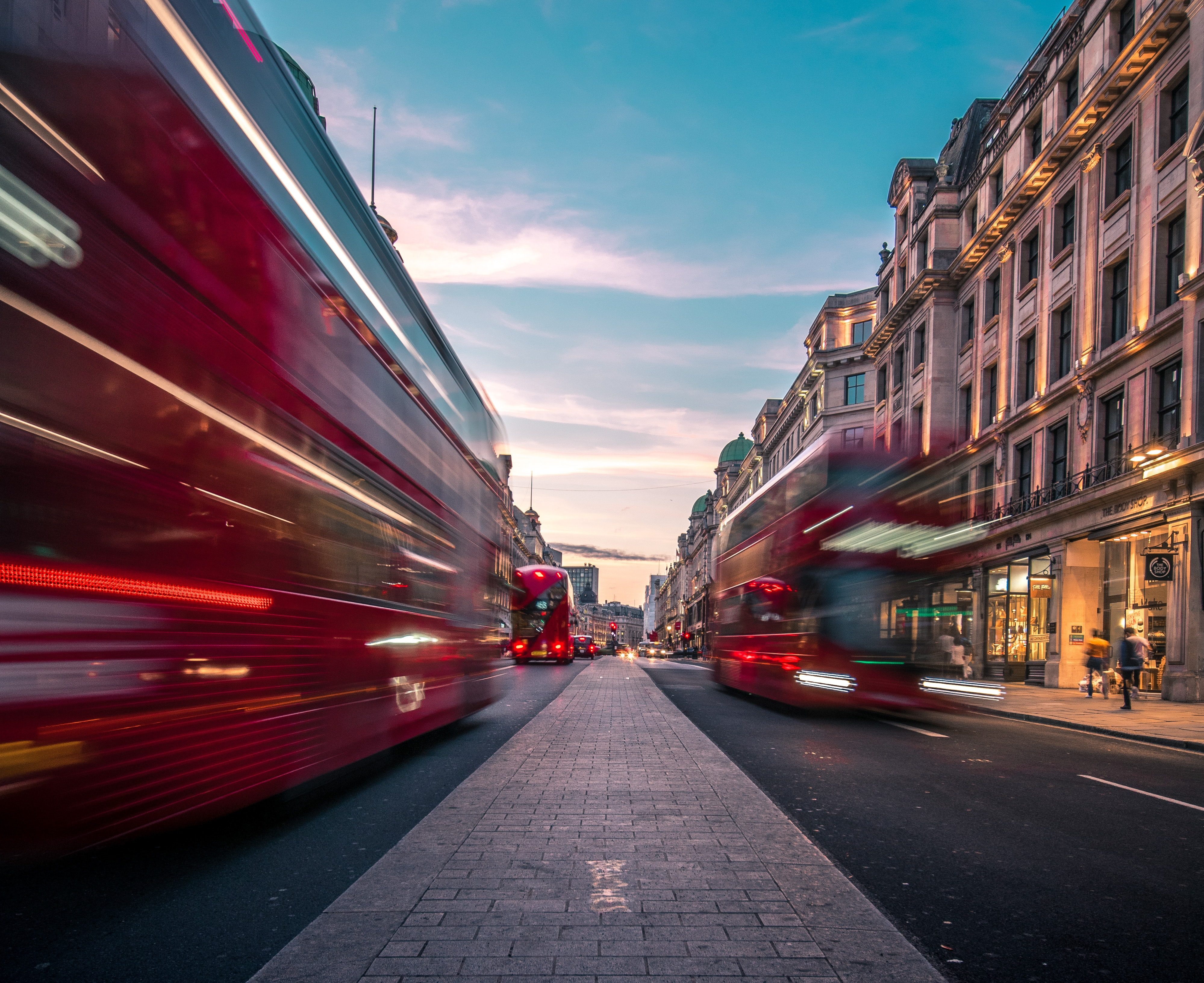 Blurred red double-decker buses move swiftly down a busy London street as dusk falls, showcasing the beauty of the city. The road is flanked by elegant buildings with shops, while the sky above is a blend of soft pink and blue hues.