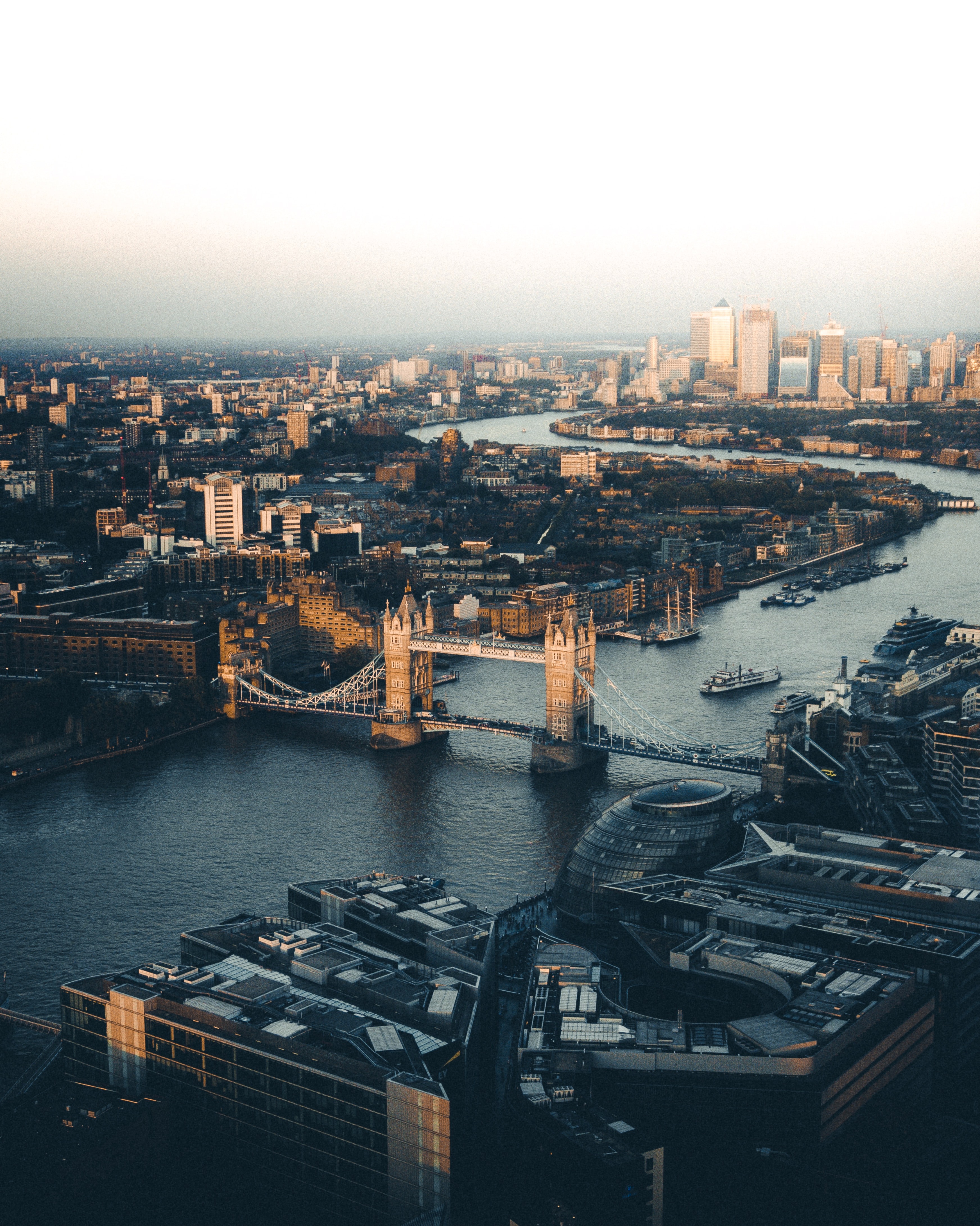 Aerial view of London featuring the iconic Tower Bridge spanning the River Thames. The city is bathed in warm light, with many buildings and skyscrapers in the background. The river weaves through the densely built urban landscape, capturing the essence of a true Londoner’s home.
