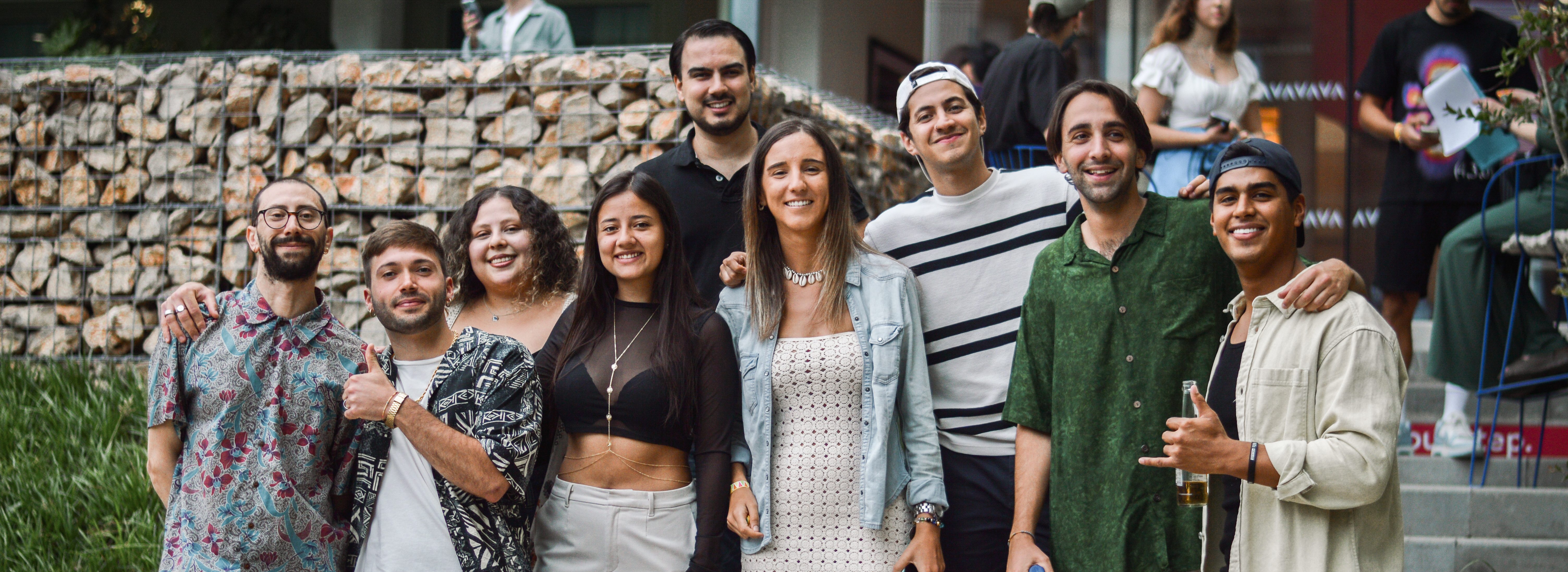 A group of nine people smiling and standing together outdoors at Vita Student Barcelona. They are casually dressed, with some holding drinks, as they prepare to enjoy the day. A stone wall and greenery add charm to the scene, while more people move around in the background.