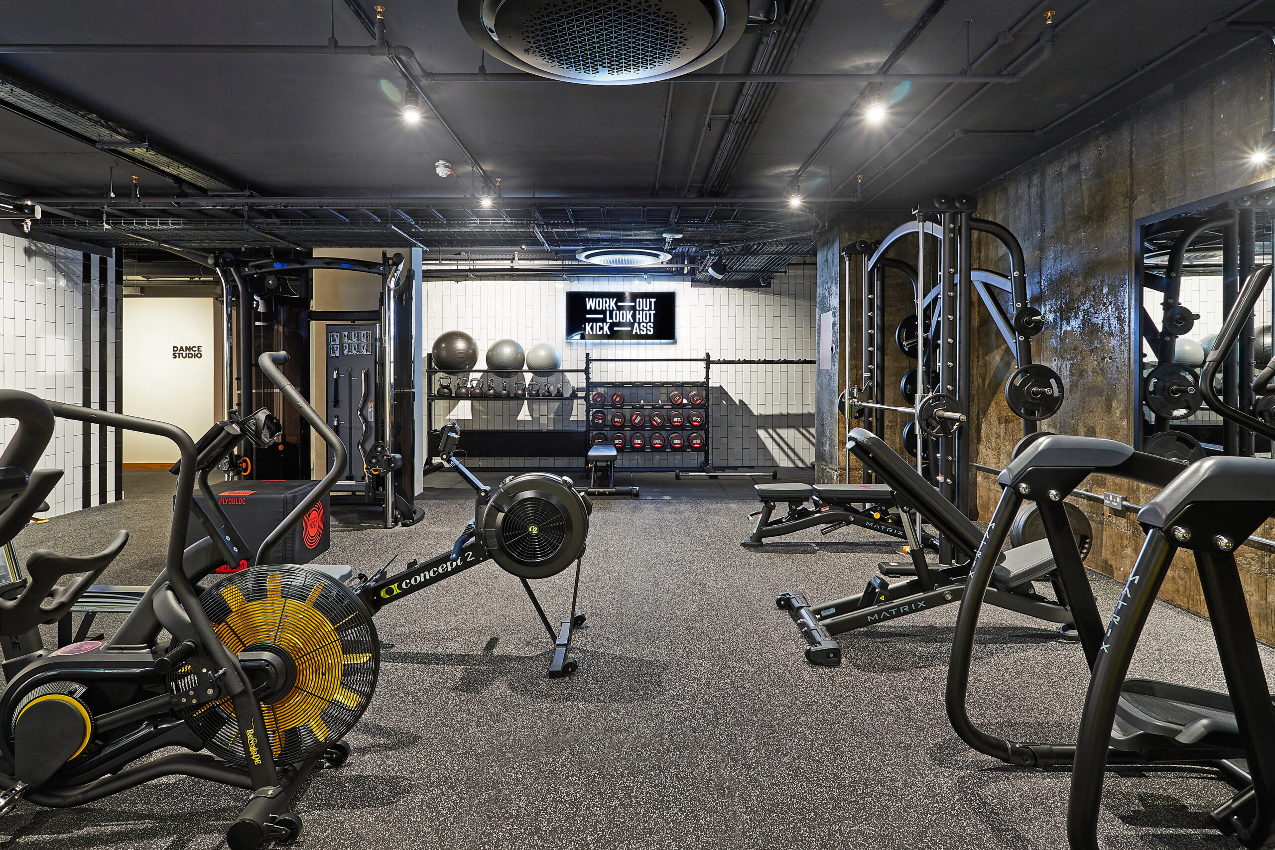Modern gym with exercise equipment including bikes, rowing machines, benches, and weights. A motivational sign on the wall reads, "Work out, suck less, kick ass." The room has a dark ceiling and bright lighting.