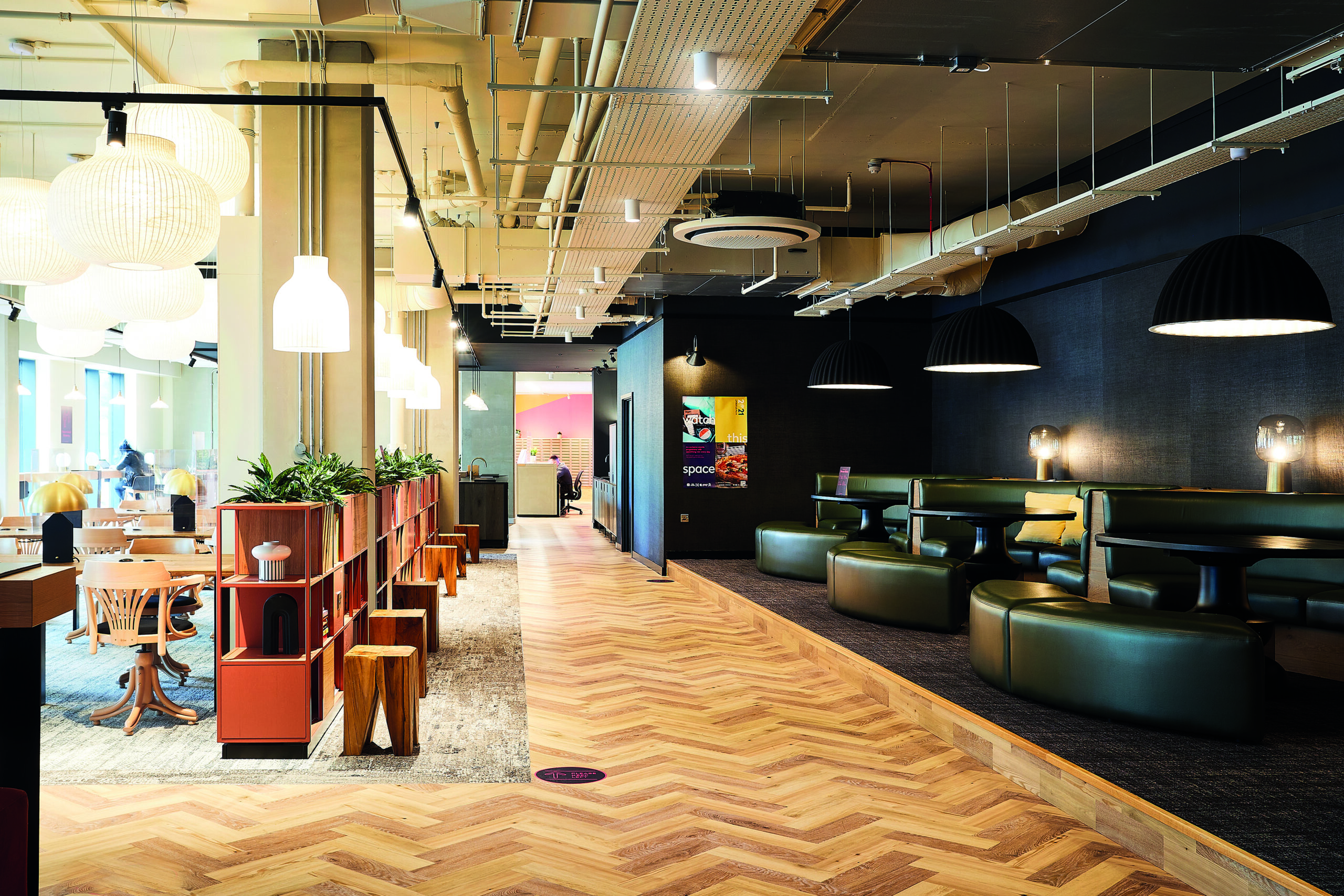 Modern office space with wooden herringbone flooring, pendant lighting, and varied seating areas, including booth-style benches. Plants and open shelving divide the space, and natural light filters through large windows on the left.