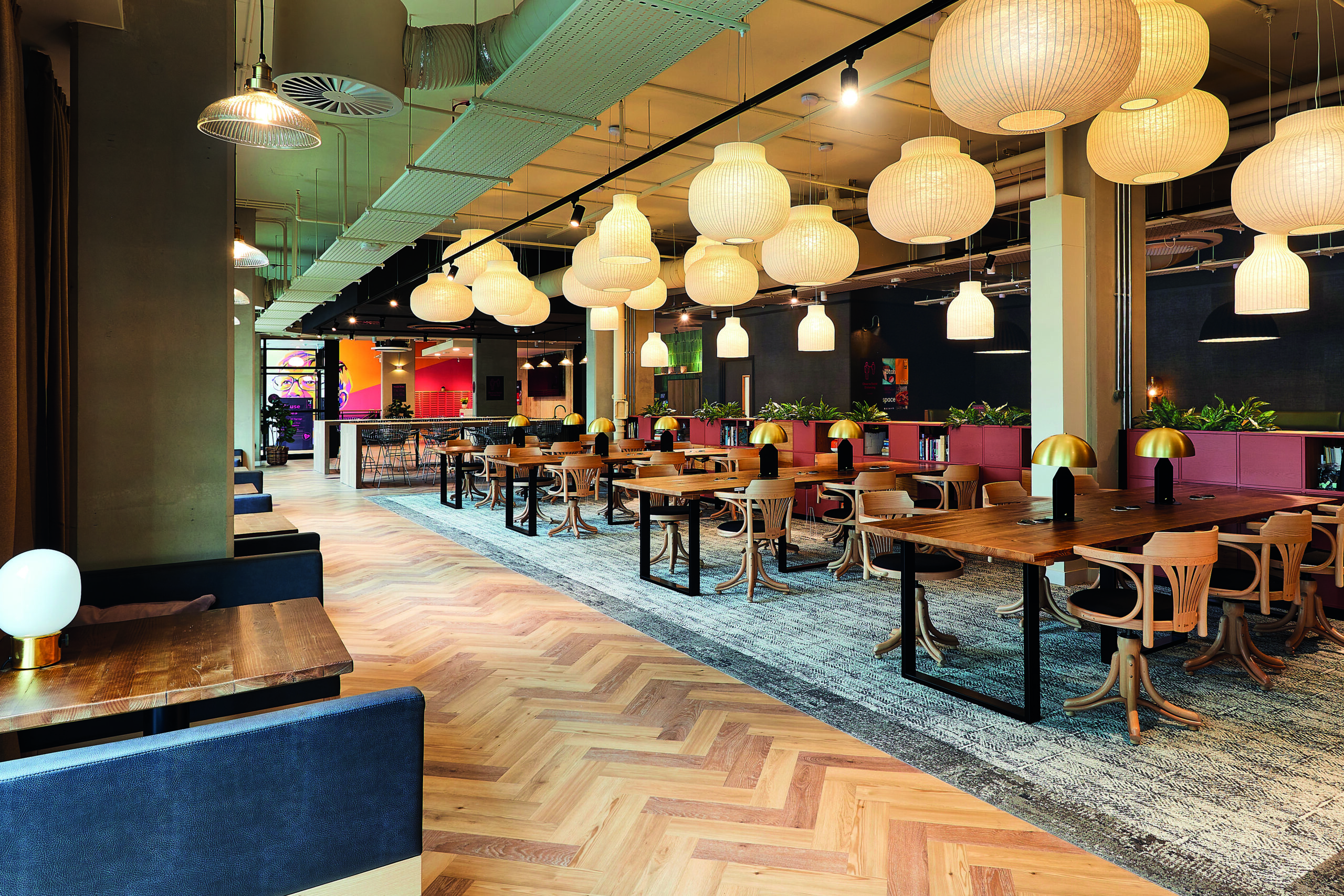 A modern restaurant interior with wooden tables and chairs, pendant lights hanging from the ceiling, and potted plants. The floor features a mix of wood and carpet, and colorful artwork is visible on the walls.