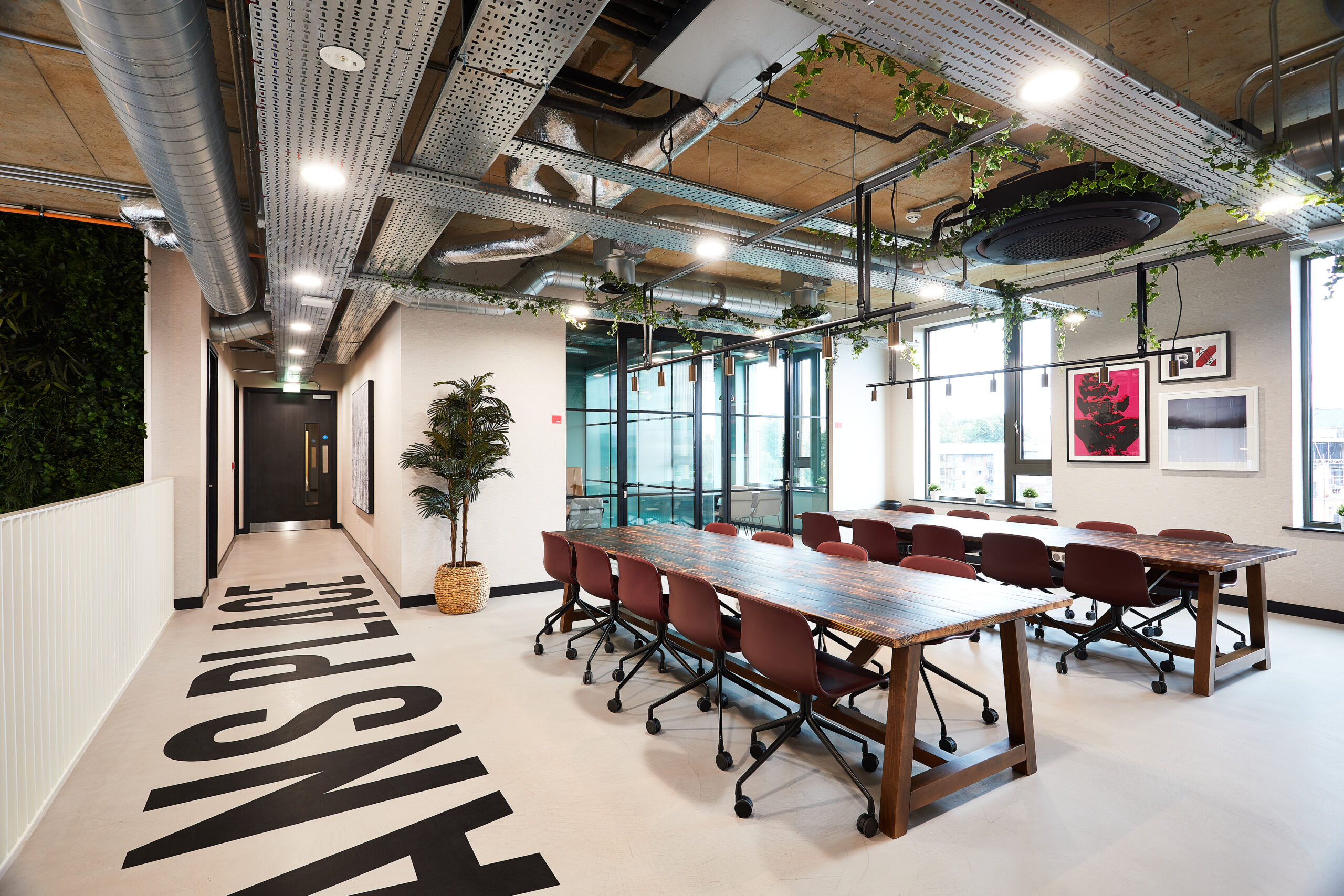 Spacious modern office with industrial design features, including exposed ducts and ceiling panels. There's a long wooden table with chairs, plants, framed art on walls, large windows, and the words "ANS PLACE" on the floor.