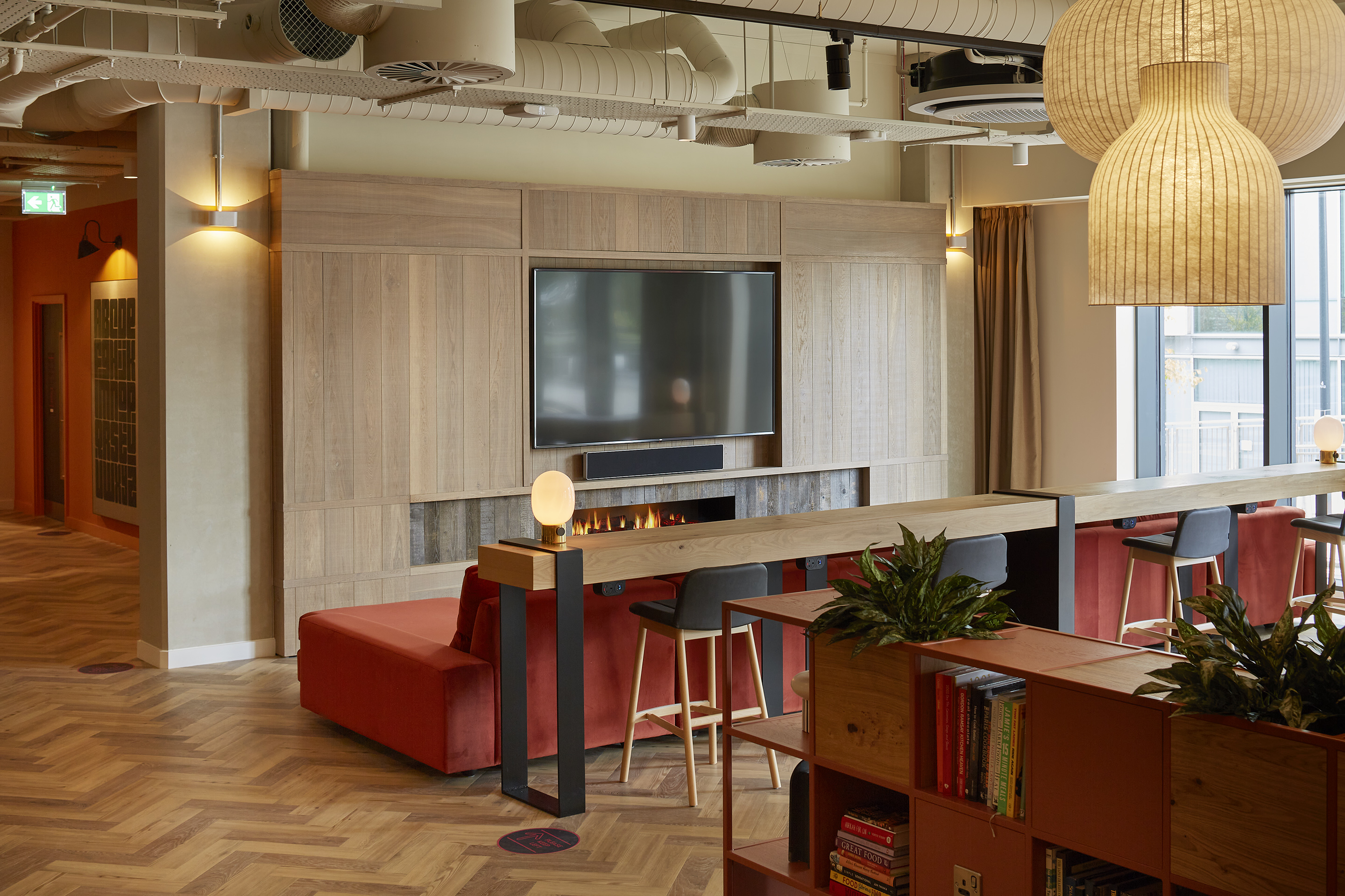 A modern lounge area with a large TV mounted above a fireplace. Red cushioned seating surrounds a wooden table with high chairs. Potted plants decorate the space, and a bookshelf filled with books adds a cozy touch. Large windows provide natural light.