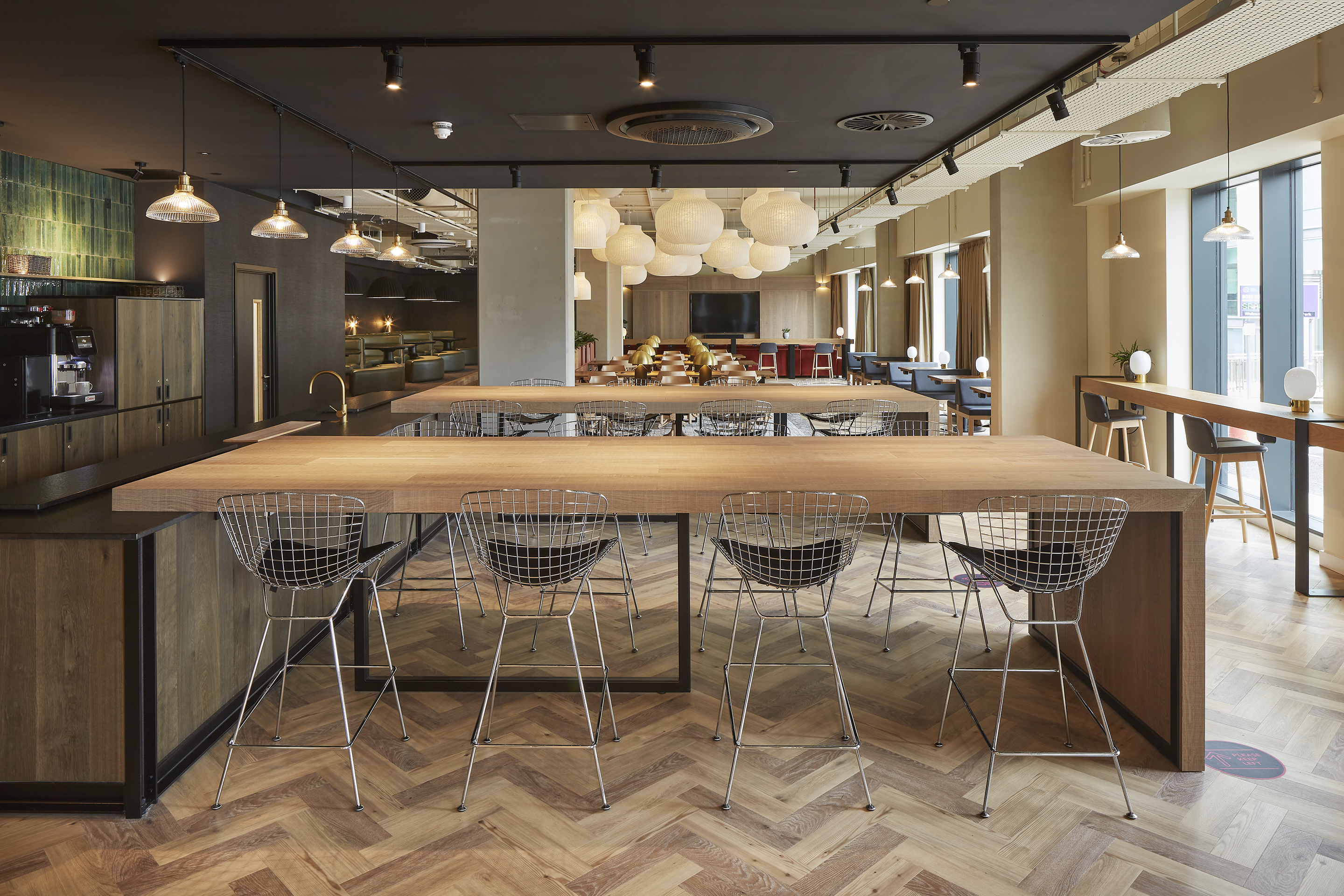 Modern cafe interior with wooden high tables and wire barstools on a herringbone parquet floor. Hanging pendant lights illuminate the space, which includes a bar area and large windows letting in natural light.