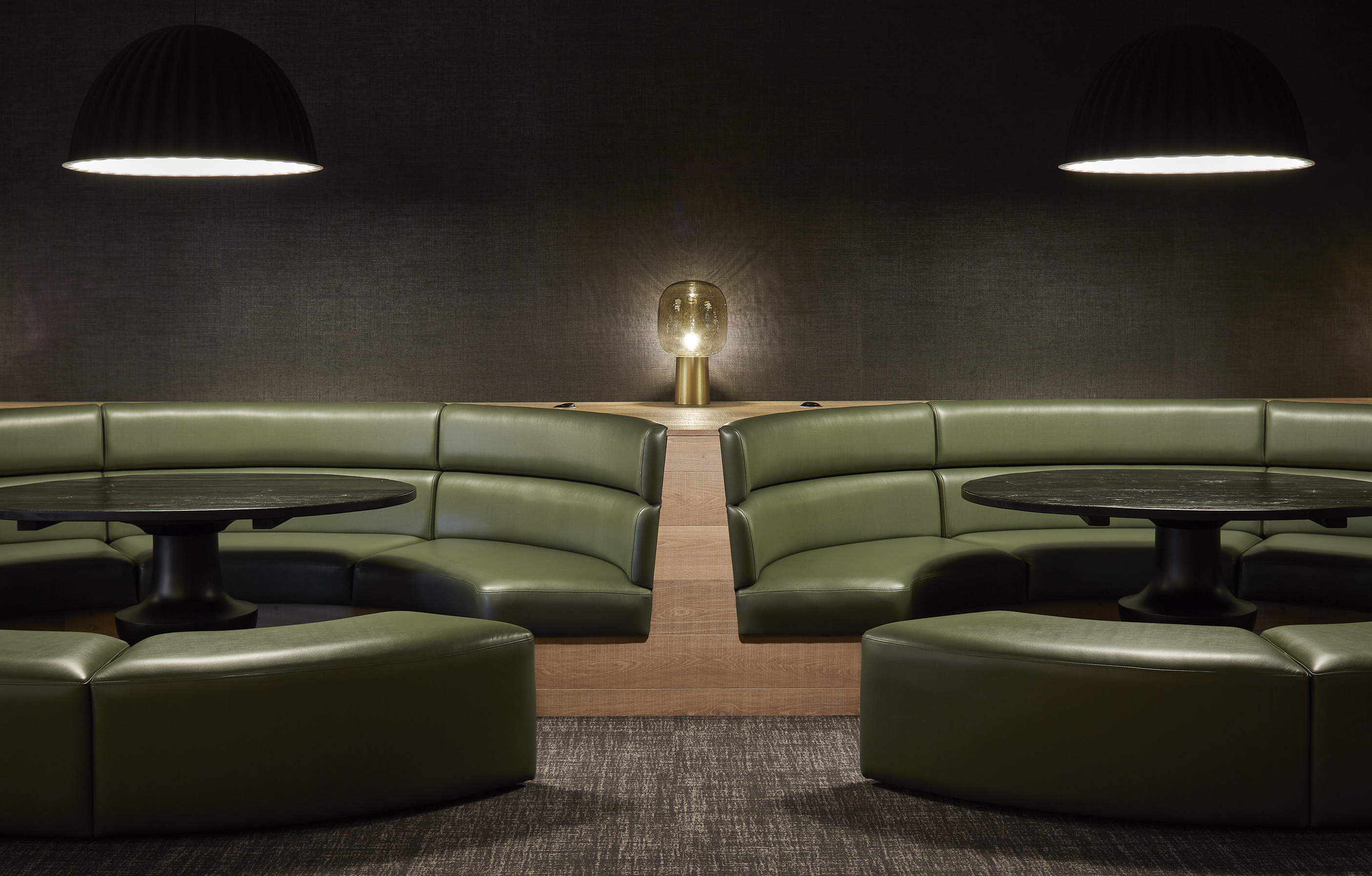 A modern, dimly-lit seating area featuring green, semi-circular couches and round black tables. Overhead, two large pendant lights hang, with a small illuminated table lamp in the center of the scene against a dark, textured backdrop.