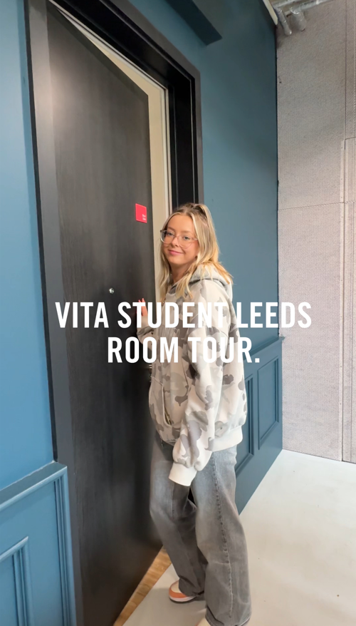 A person with long blonde hair and glasses stands in front of a dark door labeled with a red sign. They're wearing a light gray patterned jacket and jeans. Text on the image reads "Vita Student Leeds Room Tour.