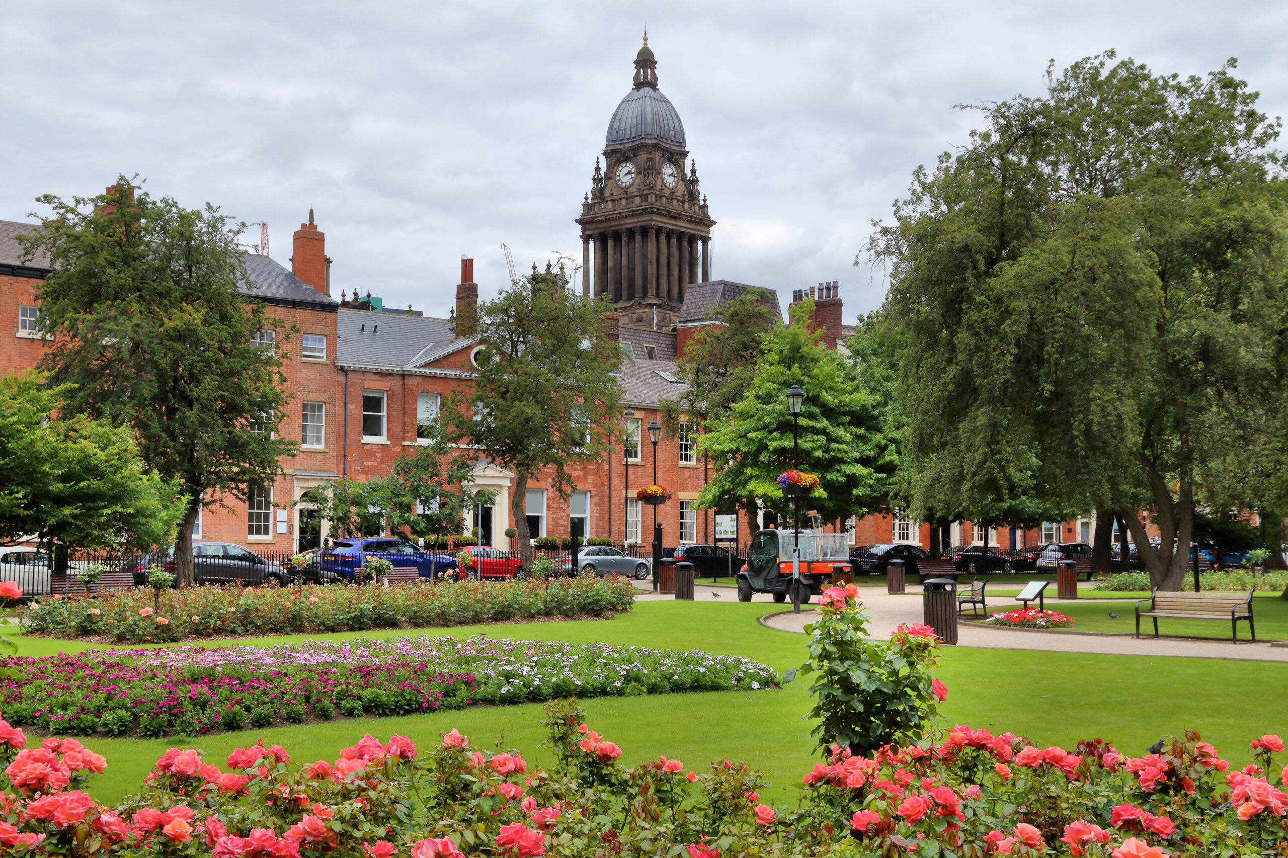 A park with vibrant red roses in the foreground, neatly trimmed grass, and flower beds. In the background are historical brick buildings and a tall clock tower. Trees line the park, and benches are scattered along the path. The sky is overcast.
