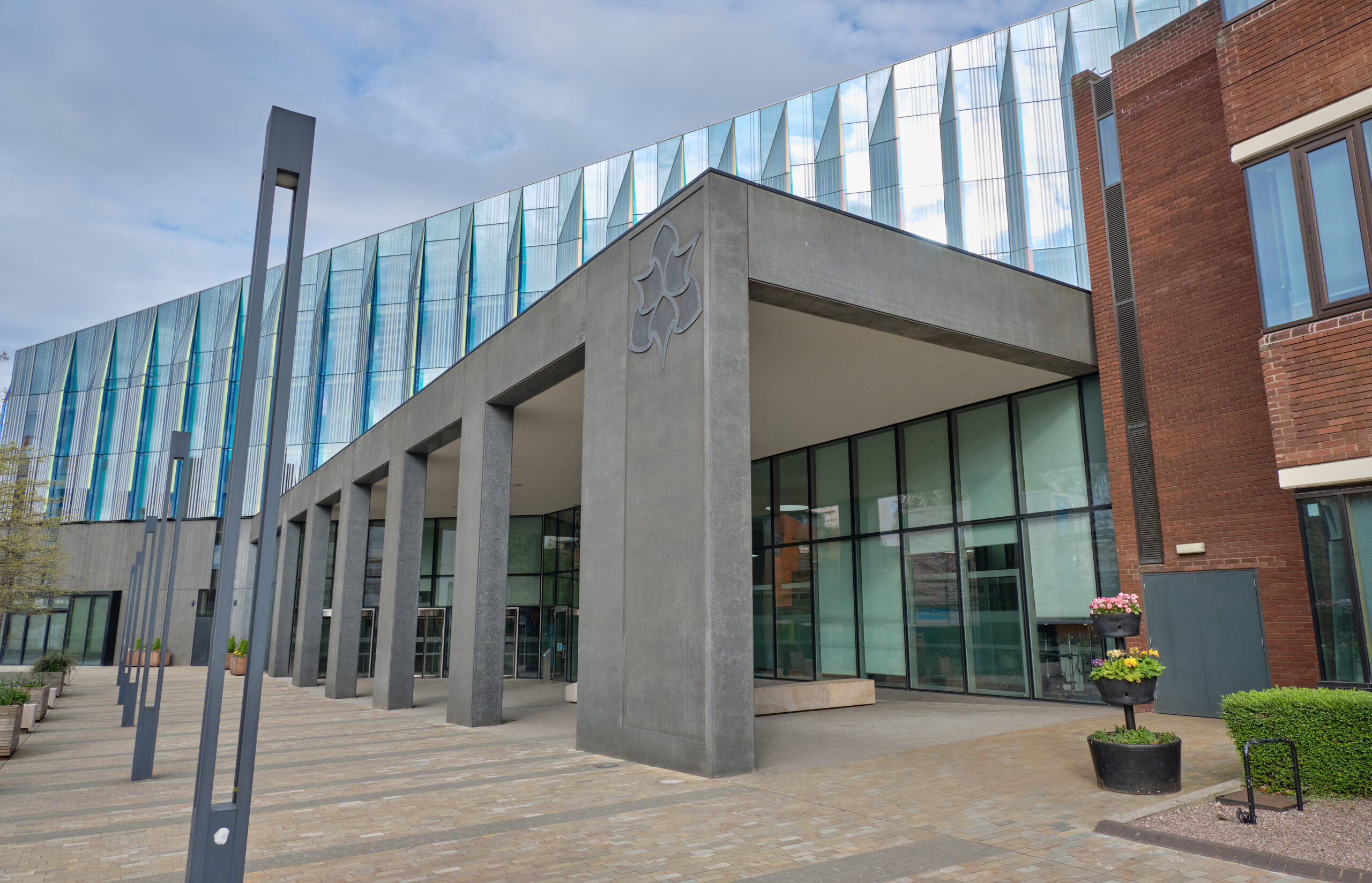 Modern building with a large glass facade and tall concrete pillars at the entrance. A geometric design is visible on the front wall. There are flower pots and benches along the paved walkway under a partly cloudy sky.