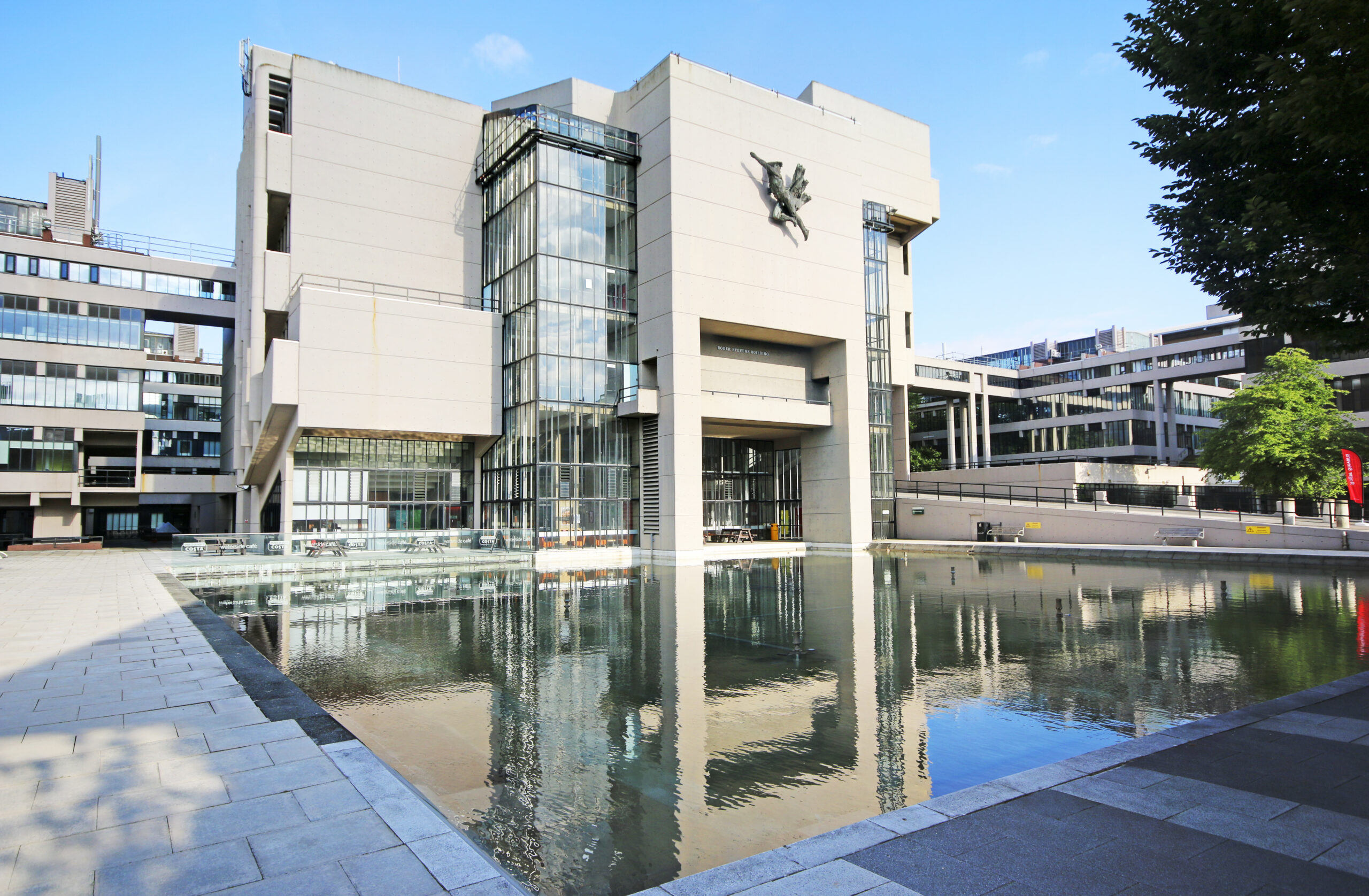 Large, modern building with geometric design and reflective glass windows set against a clear blue sky. A rectangular pool in the foreground mirrors the building. Pathways and trees surround the area, adding greenery to the scene.