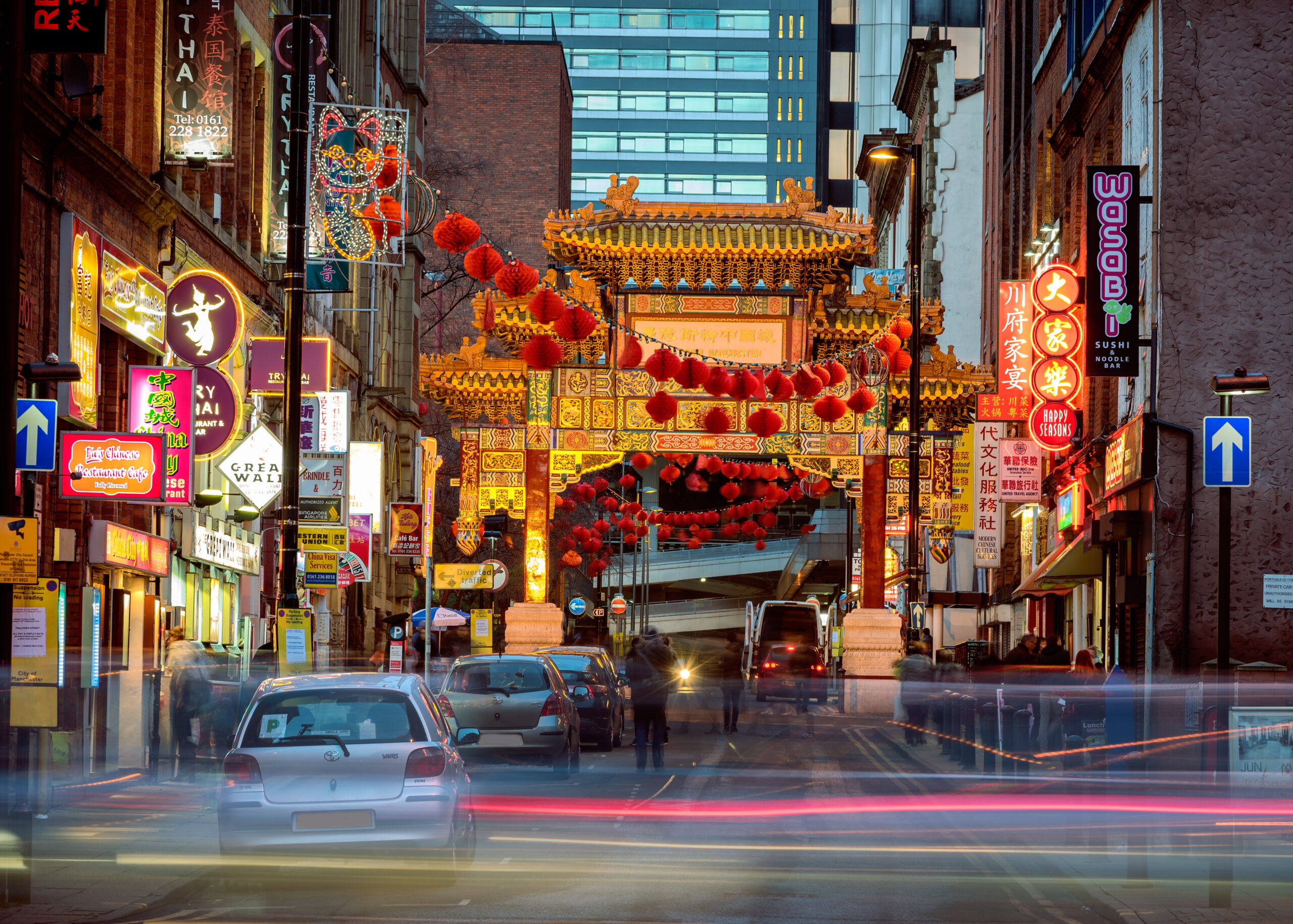 A vibrant street scene featuring an ornate archway illuminated with red lanterns. Neon signs and storefronts line the busy street, with blurred lights from passing cars. Tall buildings rise in the background, adding to the urban atmosphere.
