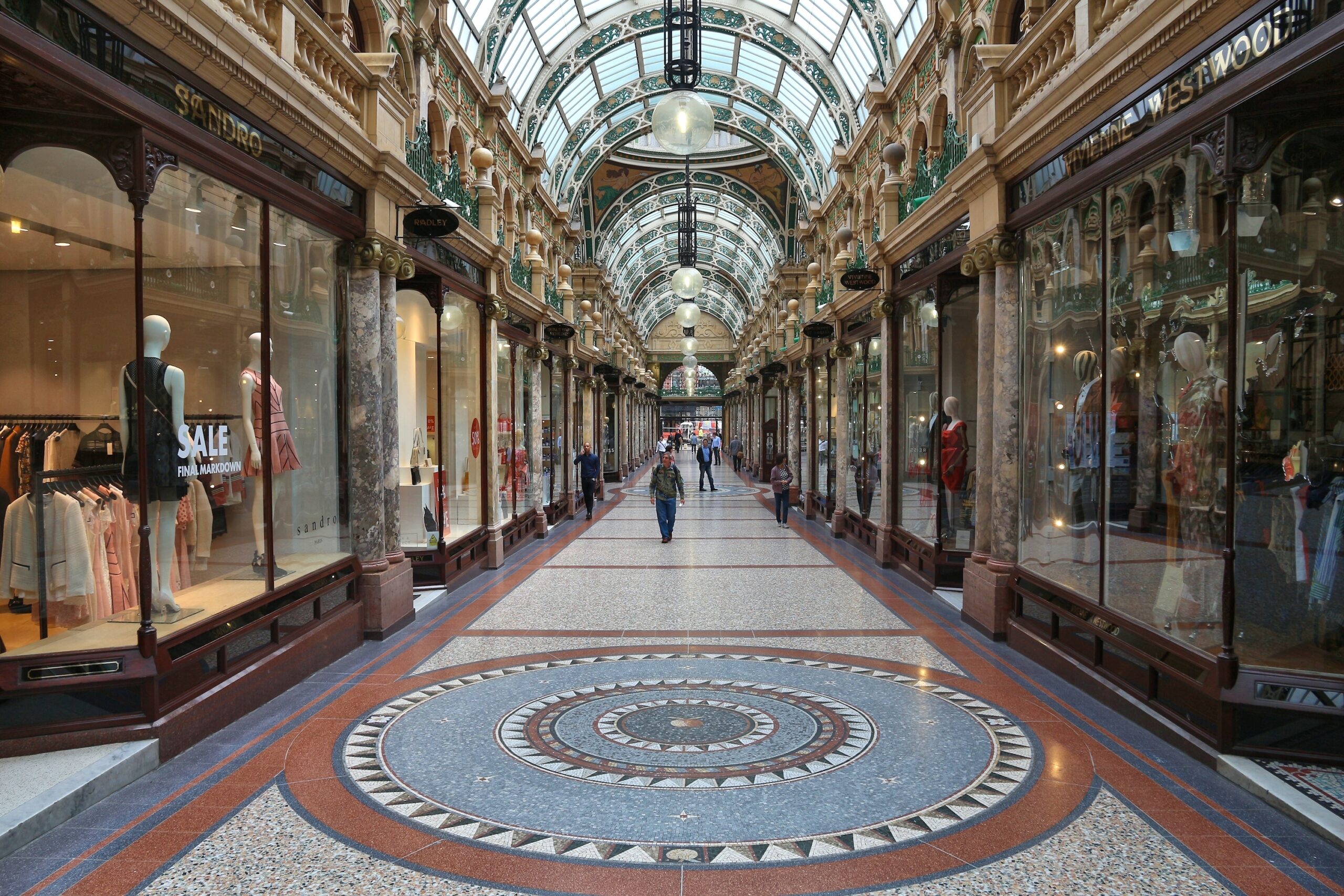 A spacious, ornate shopping arcade with a patterned mosaic floor and an arched glass ceiling. Stores with large display windows line both sides. People are walking and browsing. Elegant lighting hangs from the ceiling.