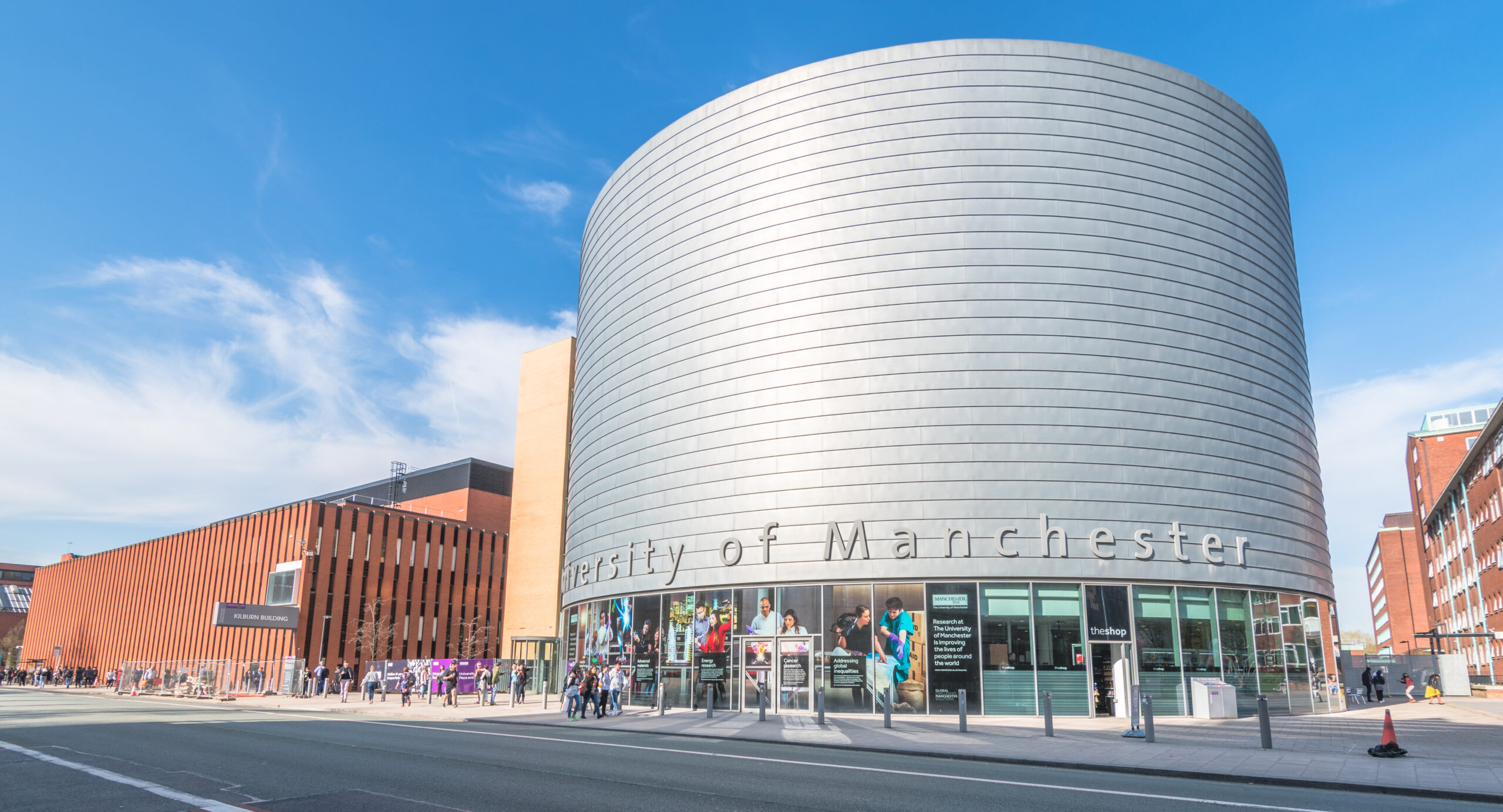 Modern building with a cylindrical silver facade and "University of Manchester" text. Clear blue sky backdrop, people walking nearby. Adjacent structure has vertical brown panels. Urban setting with street in foreground.