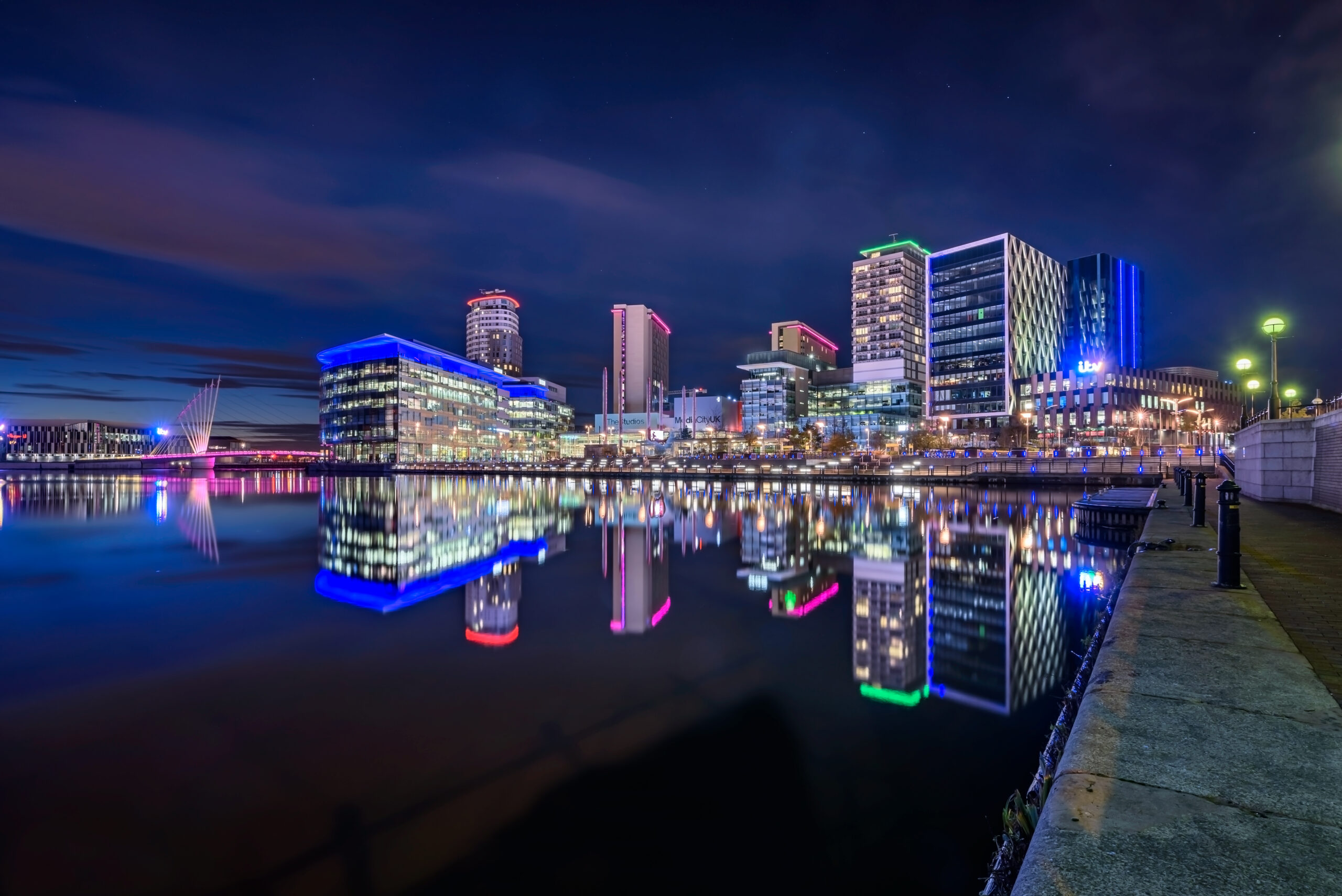 City skyline at night reflected in calm water. The buildings are illuminated with colorful lights, featuring a mix of modern architecture. The scene includes a waterfront walkway with streetlamps on the right side.