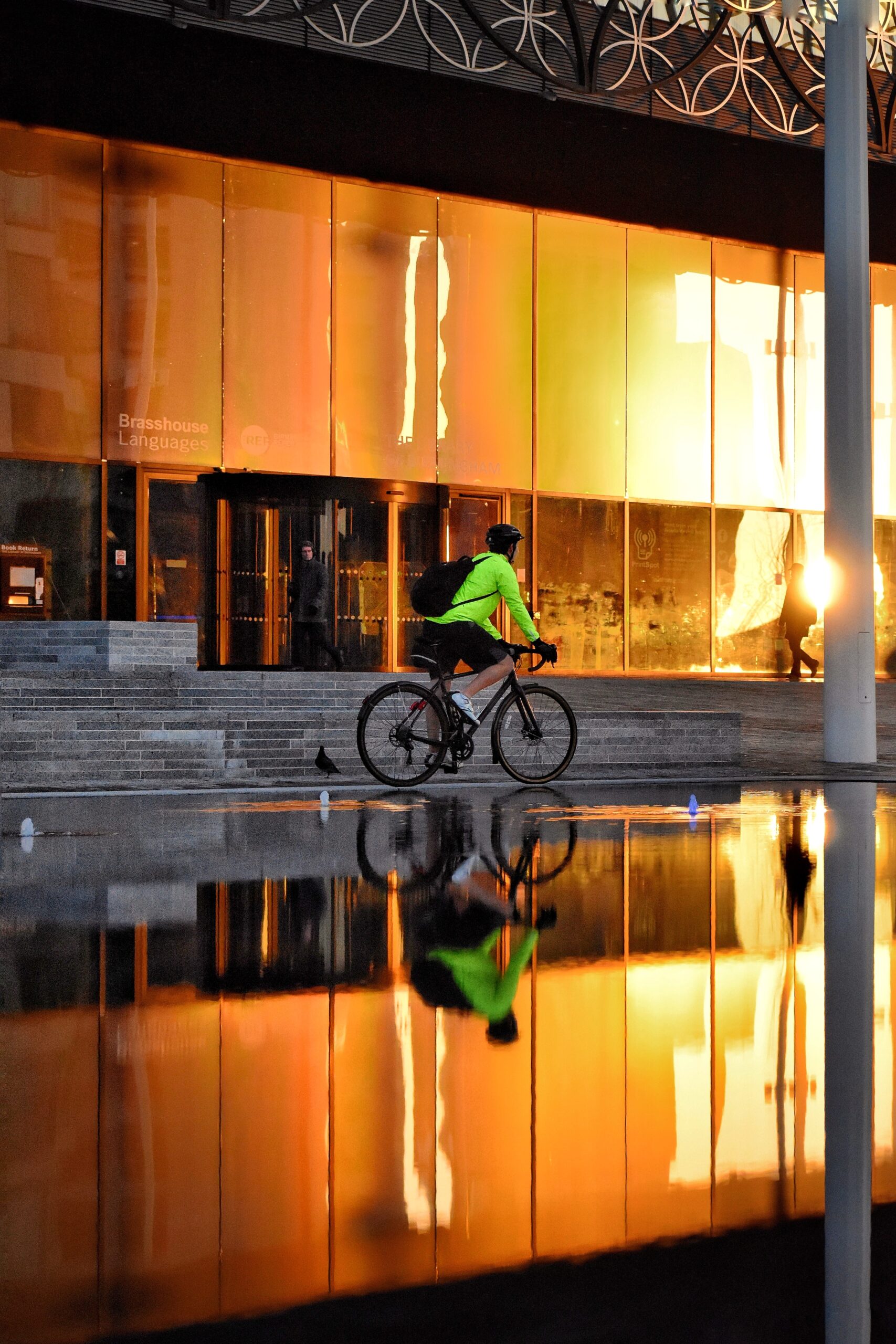Cyclist wearing a bright green jacket rides past a building with reflective orange windows. The cyclist and building are mirrored in a large puddle on the ground.