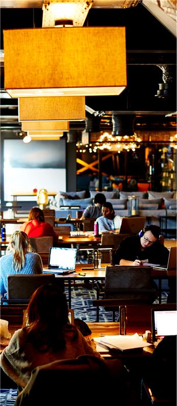 People working at laptops and writing at tables in a modern, well-lit co-working space resembling student accommodation in Birmingham, with large overhead lights and a casual, collaborative atmosphere.
