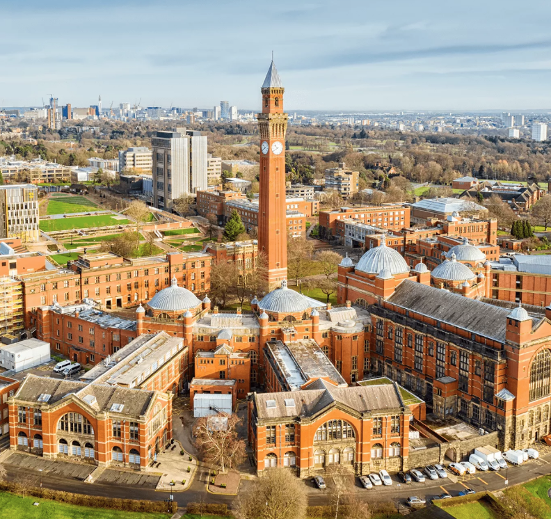 Aerial view of a large, historic university campus featuring a tall clock tower surrounded by red brick buildings with domed roofs. The cityscape spreads out in the background under a cloudy sky.