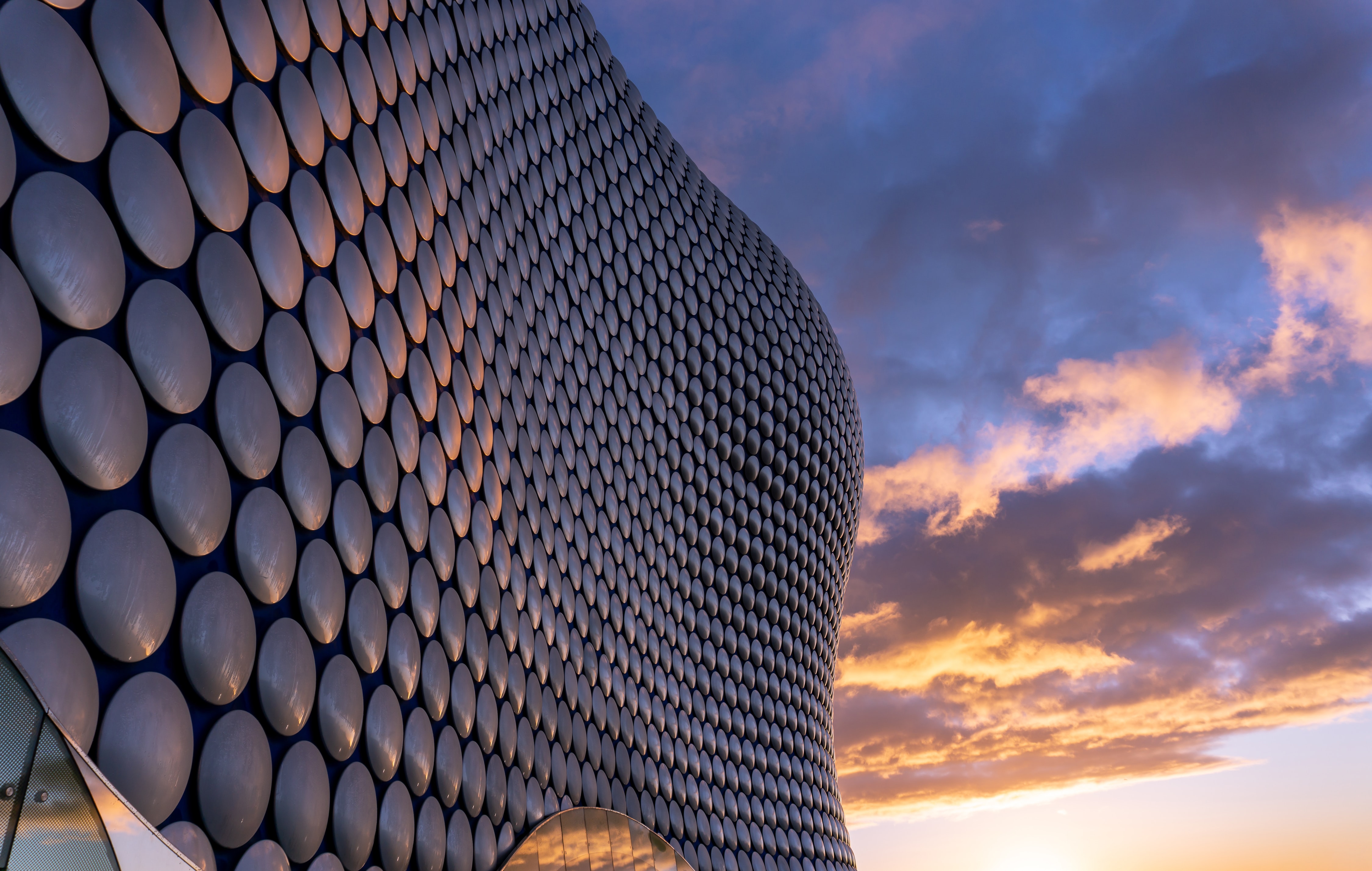 A modern architectural building with a facade of circular patterns reflects warm sunset colors against a partly cloudy sky. The building's wavy design creates a dynamic, futuristic appearance, contrasting with the natural hues of the evening.