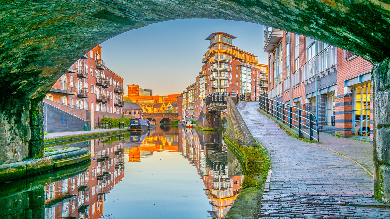 View of a canal with modern brick buildings on either side, seen through an arched stone bridge. The water reflects the colorful buildings and clear blue sky. A path runs alongside the canal, lined with a railing.