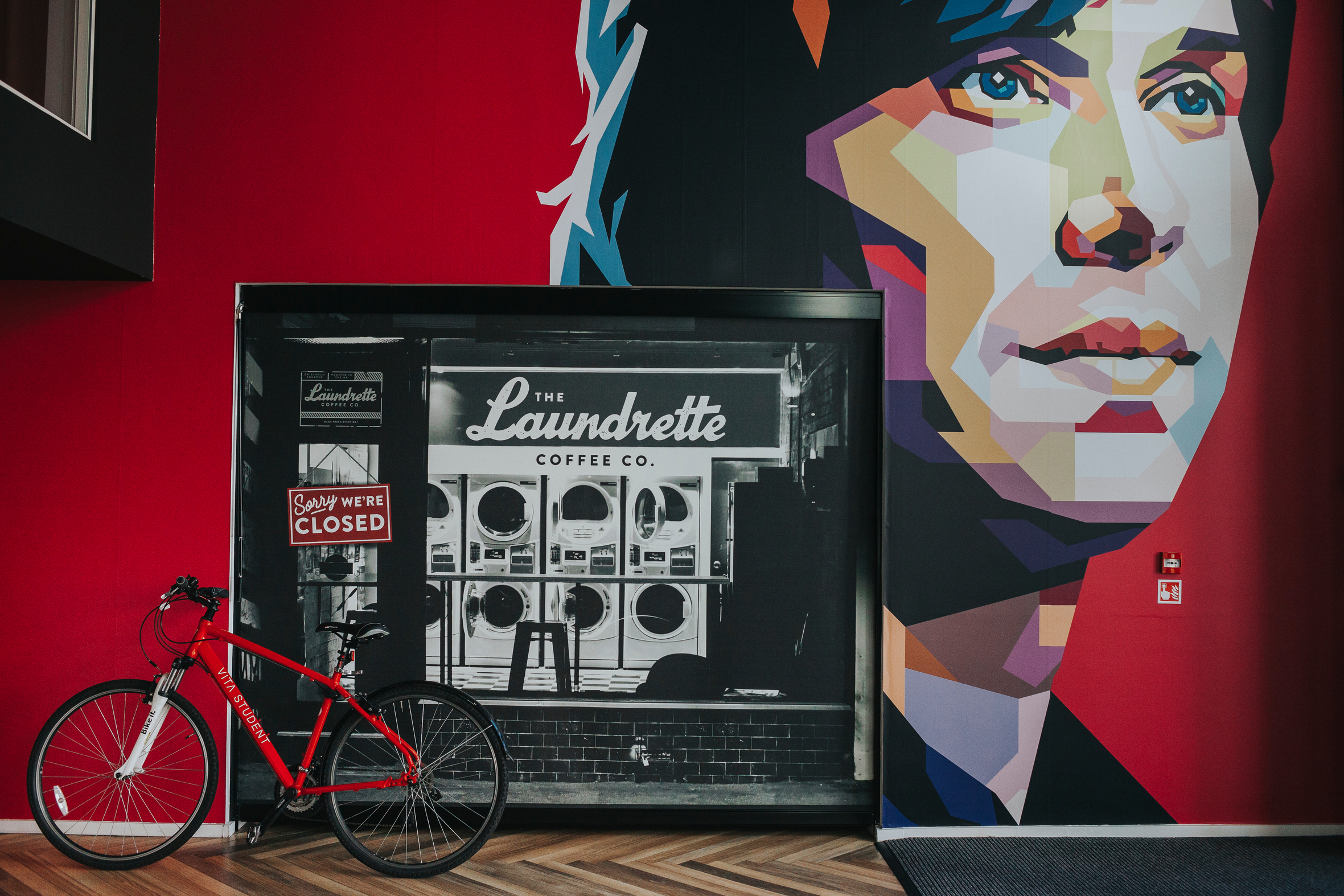 On First Street, a vibrant mural showcases a stylized portrait adjacent to a black-and-white image of a laundrette with a "Sorry, We're Closed" sign. A bright red bicycle rests in front of the artwork, while the floor below displays an intricate herringbone pattern.
