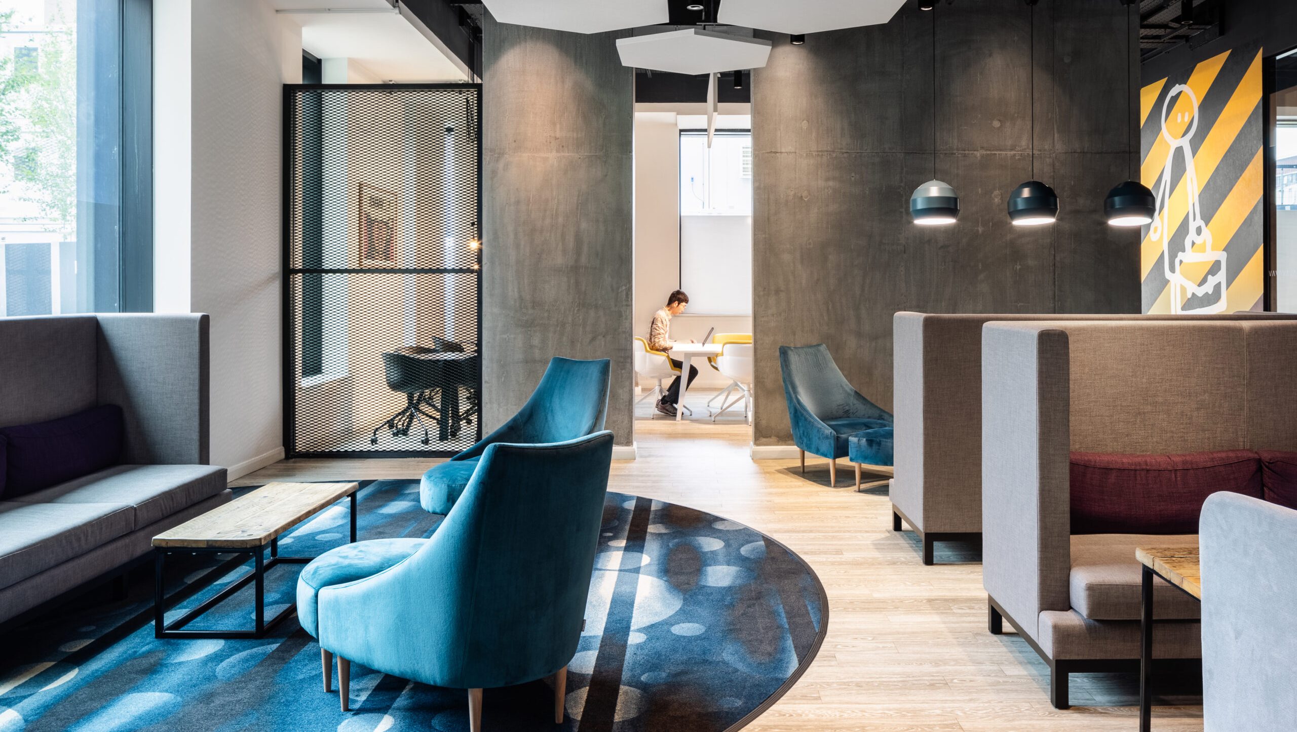 The modern First Street lounge features blue chairs and gray high-backed sofas. Wood tables rest on a patterned blue and black rug, while a person works at a desk in the background. Large wall art and pendant lights enhance the contemporary vibe.