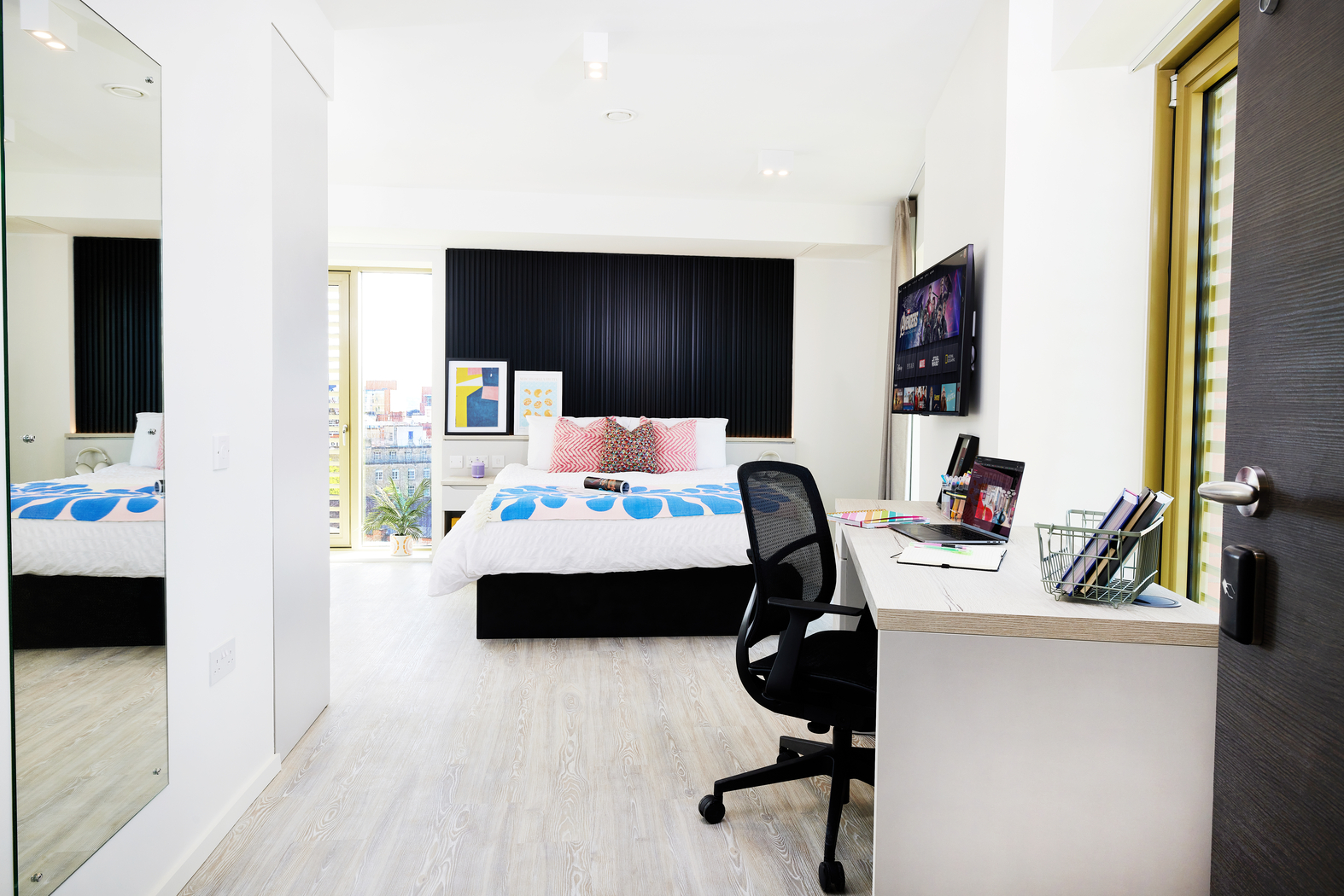 Located on Bruce Street, this modern bedroom features a bed with blue and white bedding. A work desk and chair sit in the foreground, equipped with a computer screen, office supplies, and a window offering natural light. Minimalist decor is highlighted by a mirror on the wall.