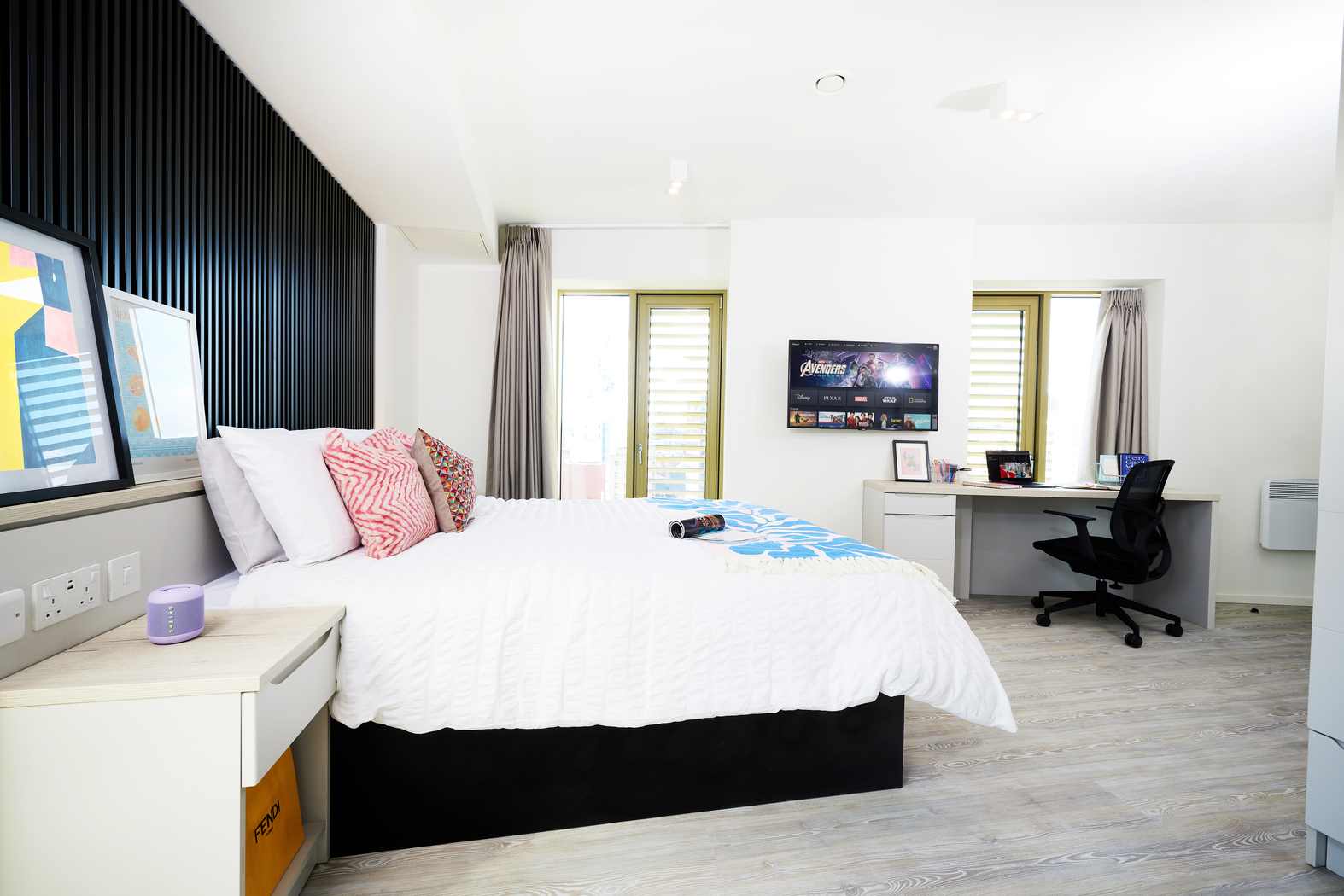 Bright, modern bedroom on Bruce Street with a large bed adorned with colorful pillows on the left. A TV is mounted on a wall, and a desk with a chair is on the right. Lots of natural light flows in from windows at the back.