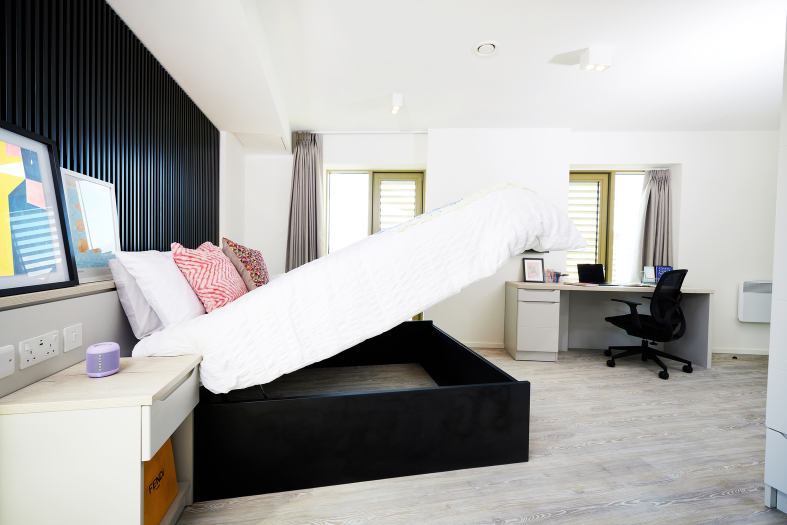 A bright bedroom on Bruce Street features a black bed frame with hidden storage, holding a white duvet and pink pillows. The room includes a desk with a chair, a computer monitor, and a painting on the wall. Two windows provide natural light.
