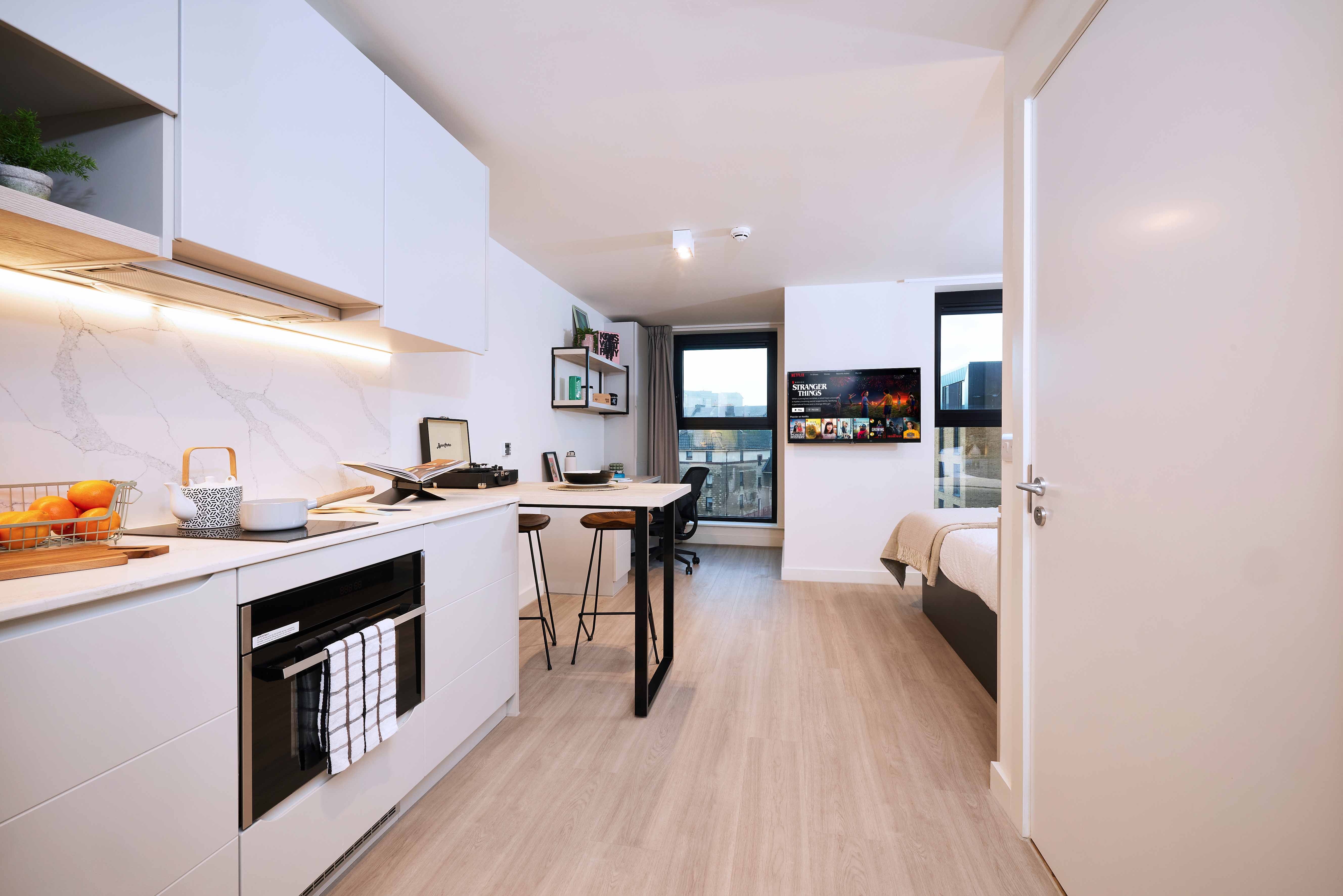 Modern studio apartment with light wood flooring, featuring a compact kitchen with white cabinets and a marble backsplash. A small dining area with two stools is adjacent, and a bed and TV are in the background near a window with a city view.