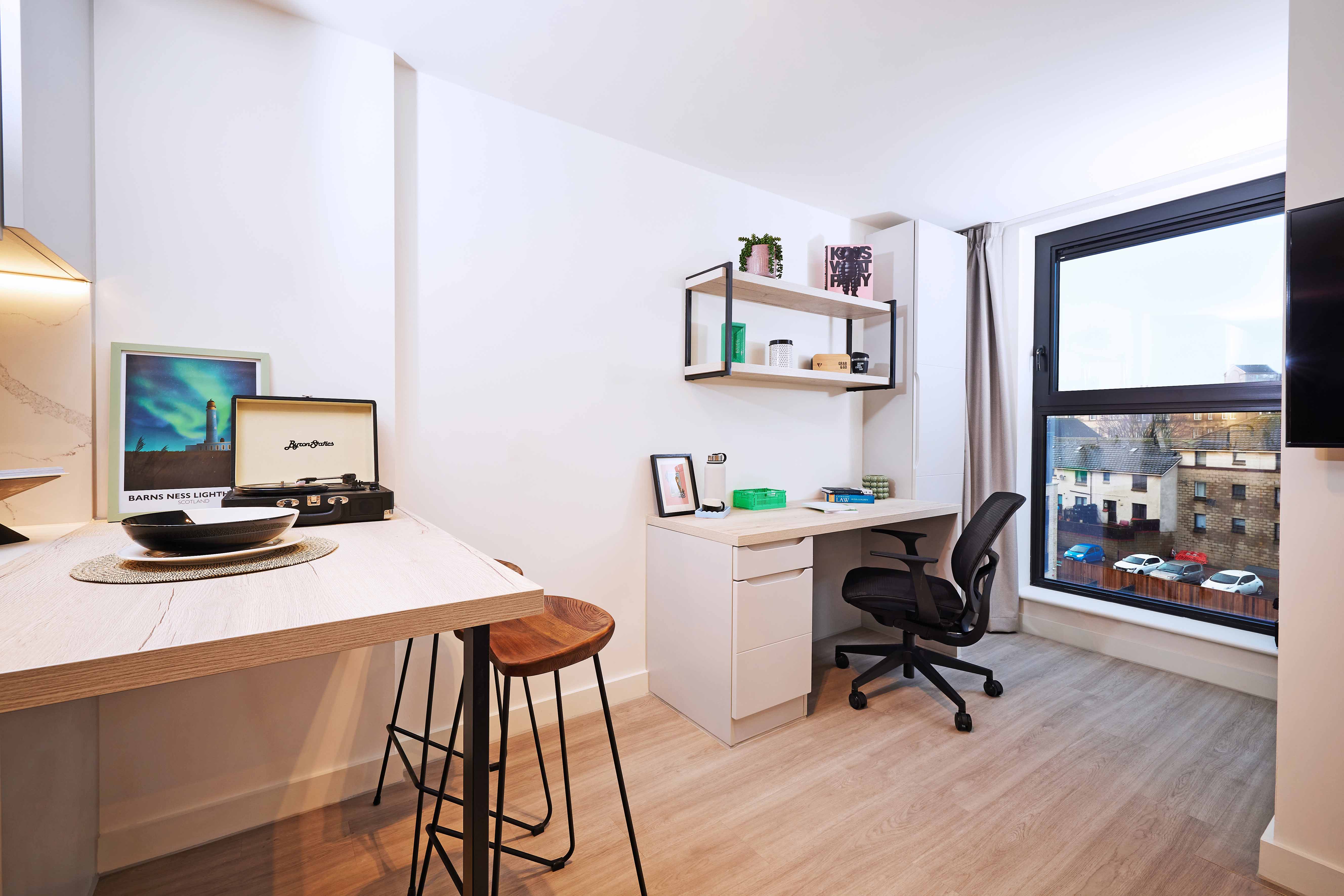 A modern home office with a desk, ergonomic chair, shelves with books and plants, and a large window displaying a city view. A record player and framed pictures are on an adjacent wooden counter, with tall stools tucked underneath.