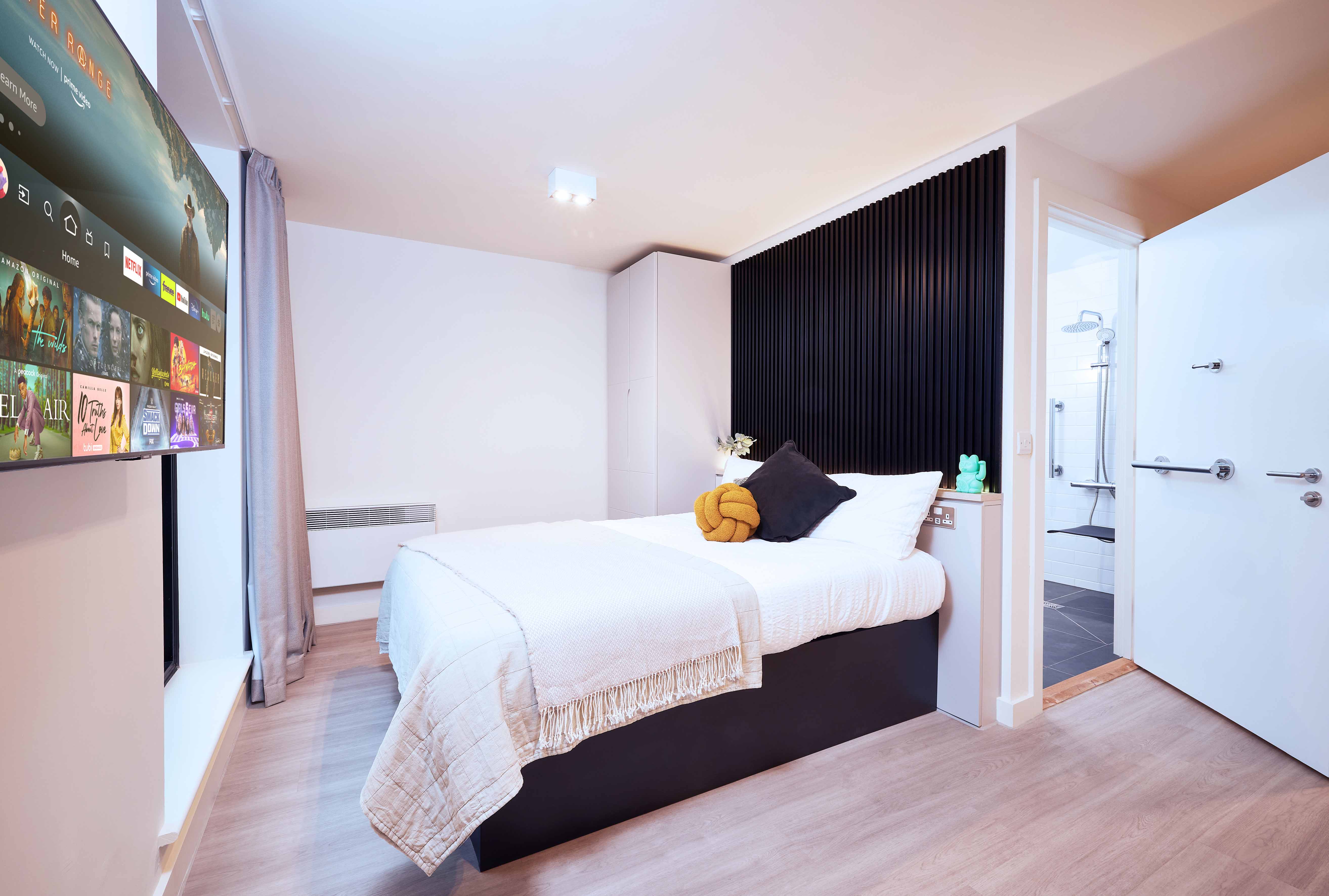 A modern bedroom featuring a bed with white linens and a mustard pillow, a wall-mounted TV displaying a streaming service menu, and an open door leading to a bathroom. The room has wooden flooring and minimalist decor.
