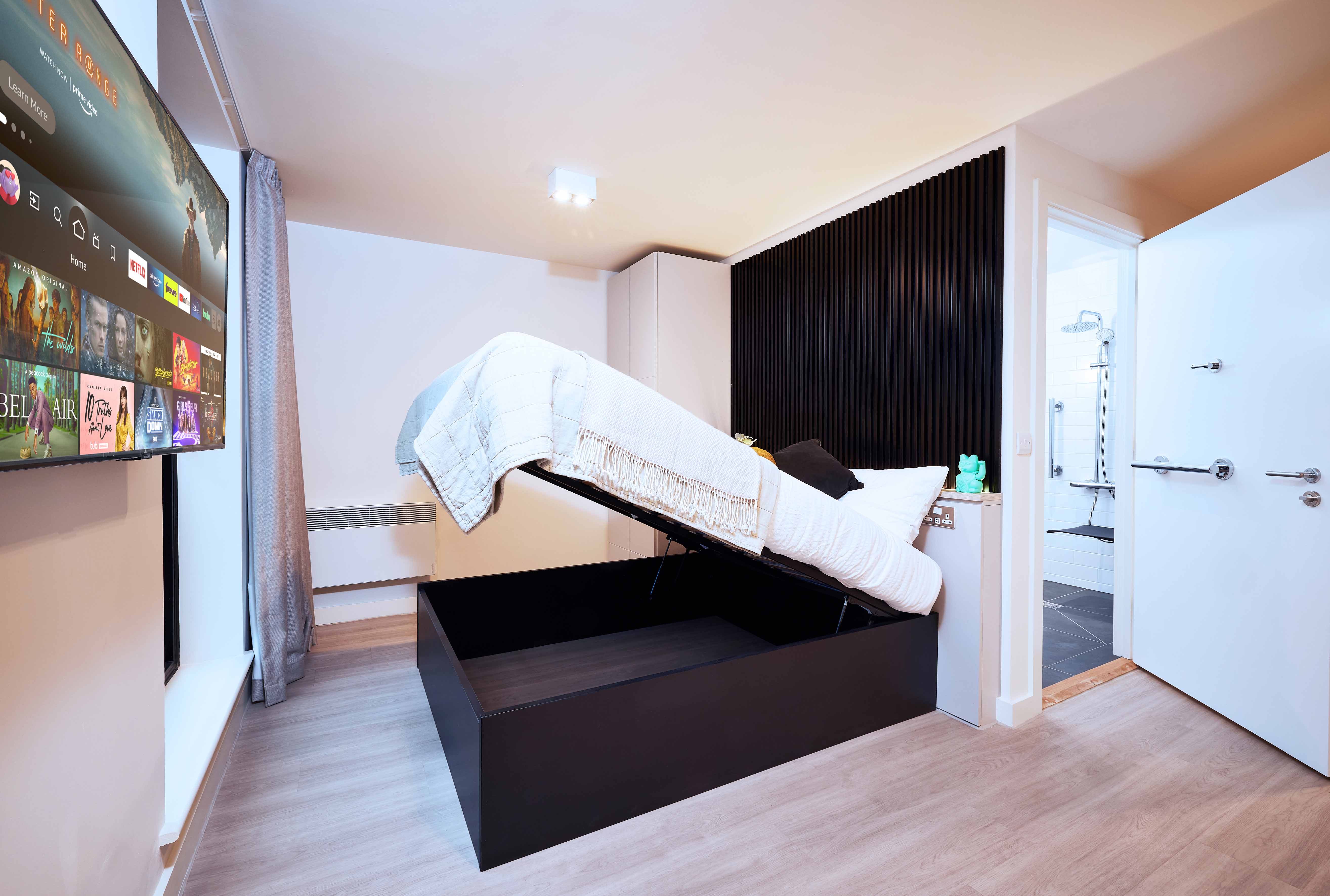 Modern bedroom with a bed that lifts to reveal storage underneath. A TV on the wall displays streaming options. The room has light wood flooring, white walls, and an open door leading to a bathroom. Grey curtains hang beside a large window.