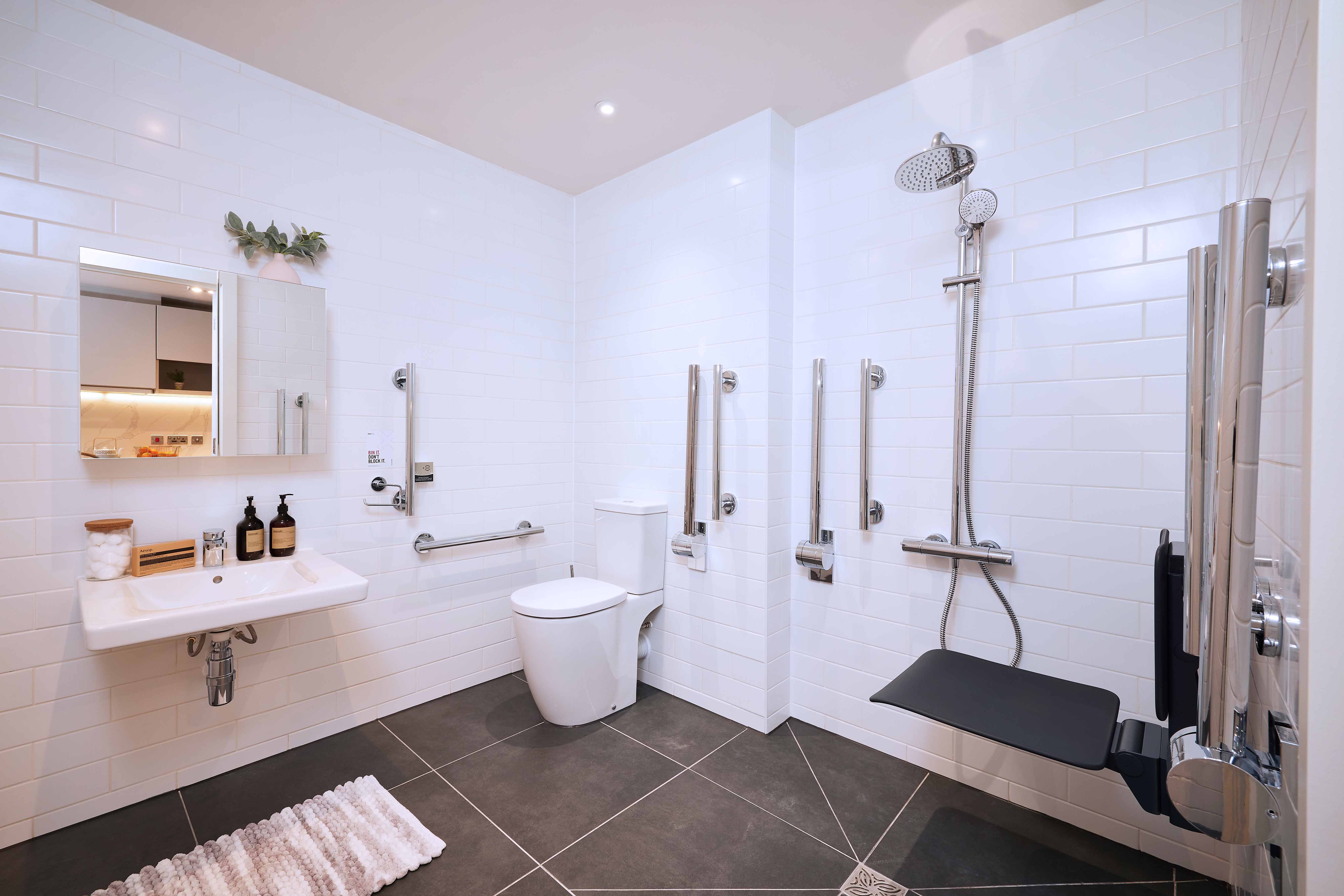 Accessible bathroom with white tiles, featuring a wall-mounted sink, grab bars, a shower with a foldable seat, and a toilet. Shelves with accessories are above the sink, and the floor is dark tile with a plush rug.