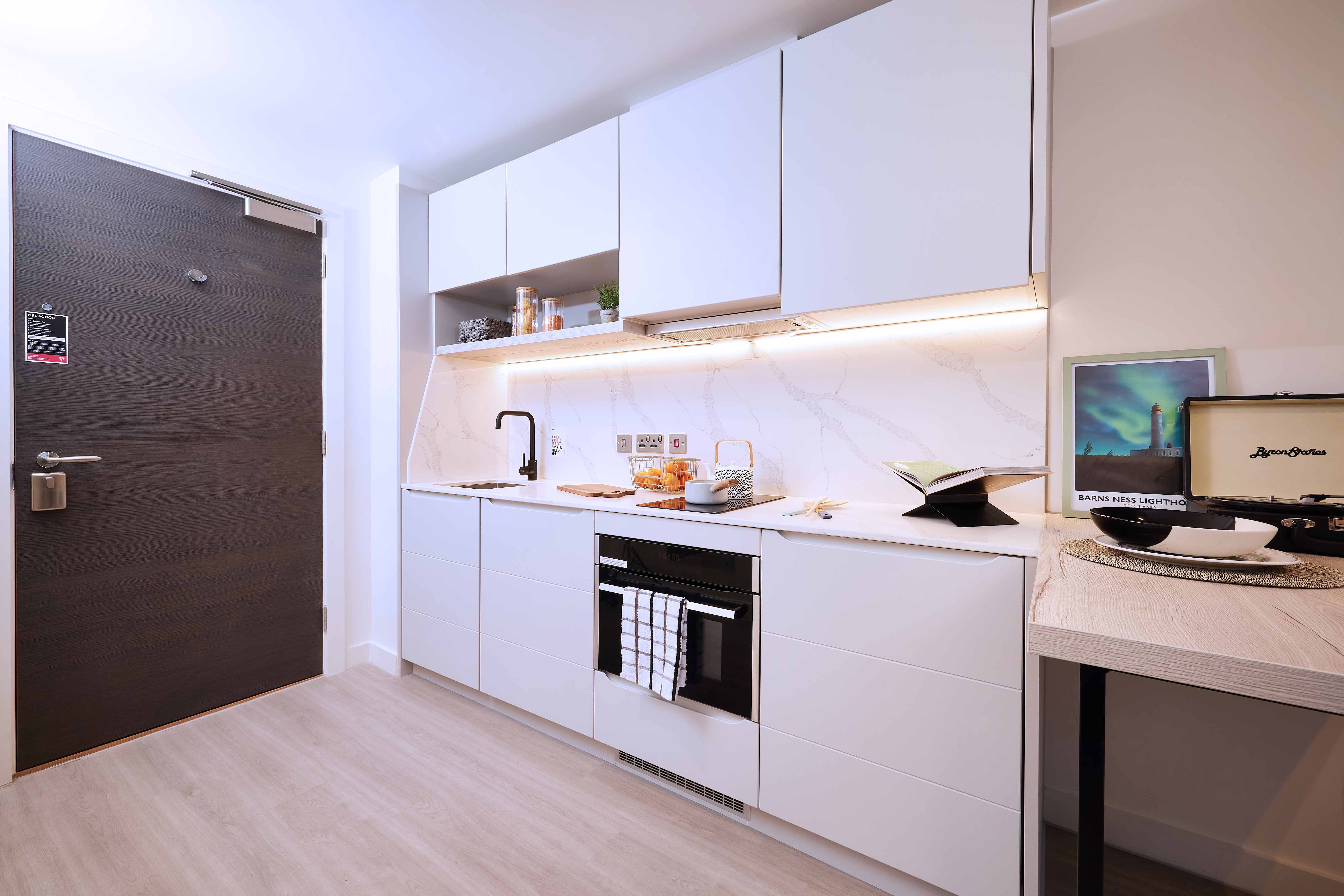 Modern kitchen with white cabinets, marble backsplash, and light wood flooring. Features a built-in oven, sink, and countertop decor. A table with books and decor is on the right near a dark wooden door. Bright and minimalist design.