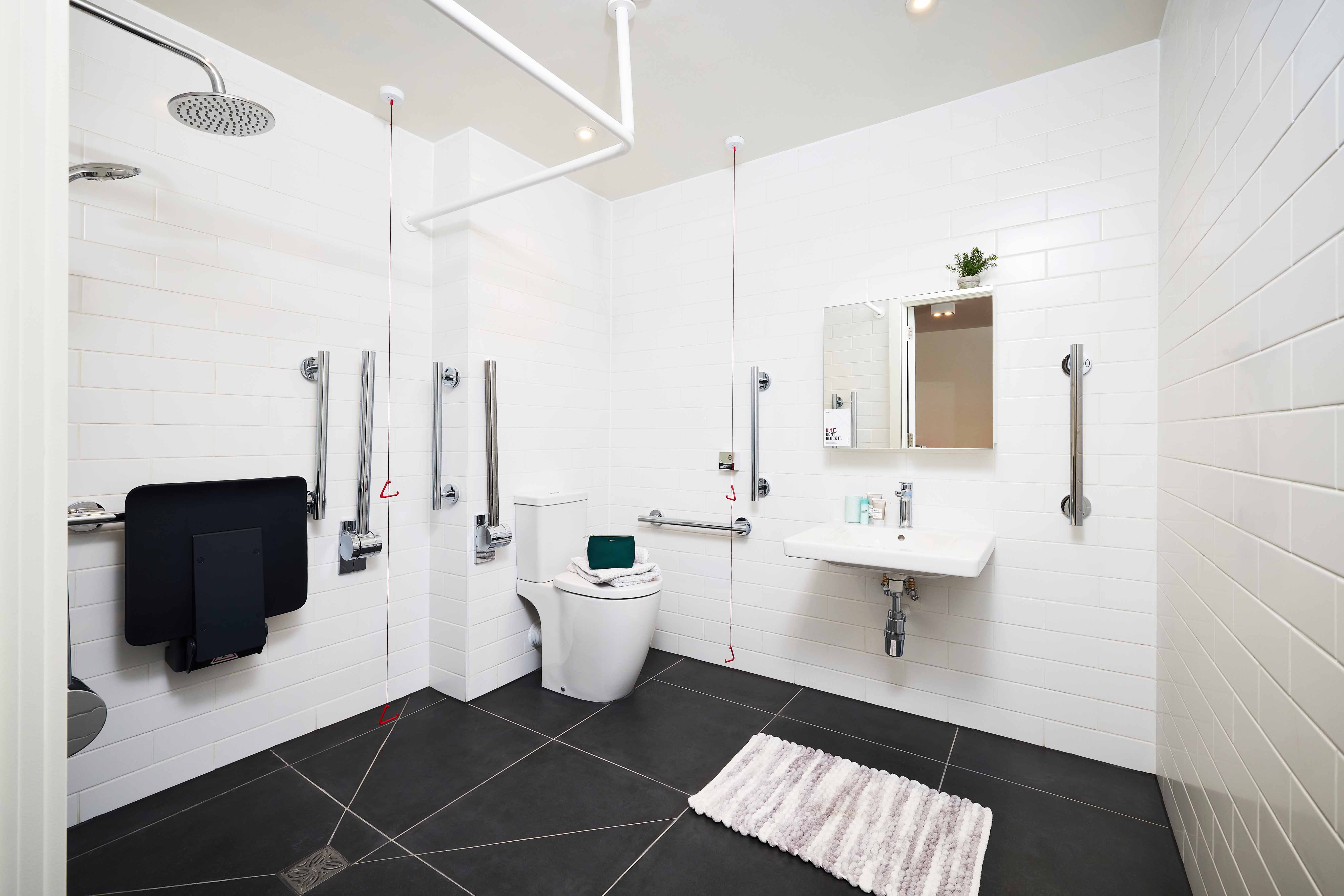 Spacious, accessible bathroom with white tiled walls and dark floor. Features include a wall-mounted toilet, grab bars, a foldable shower seat, overhead shower, sink with mirror, and small potted plant on the shelf. A striped rug lies on the floor.