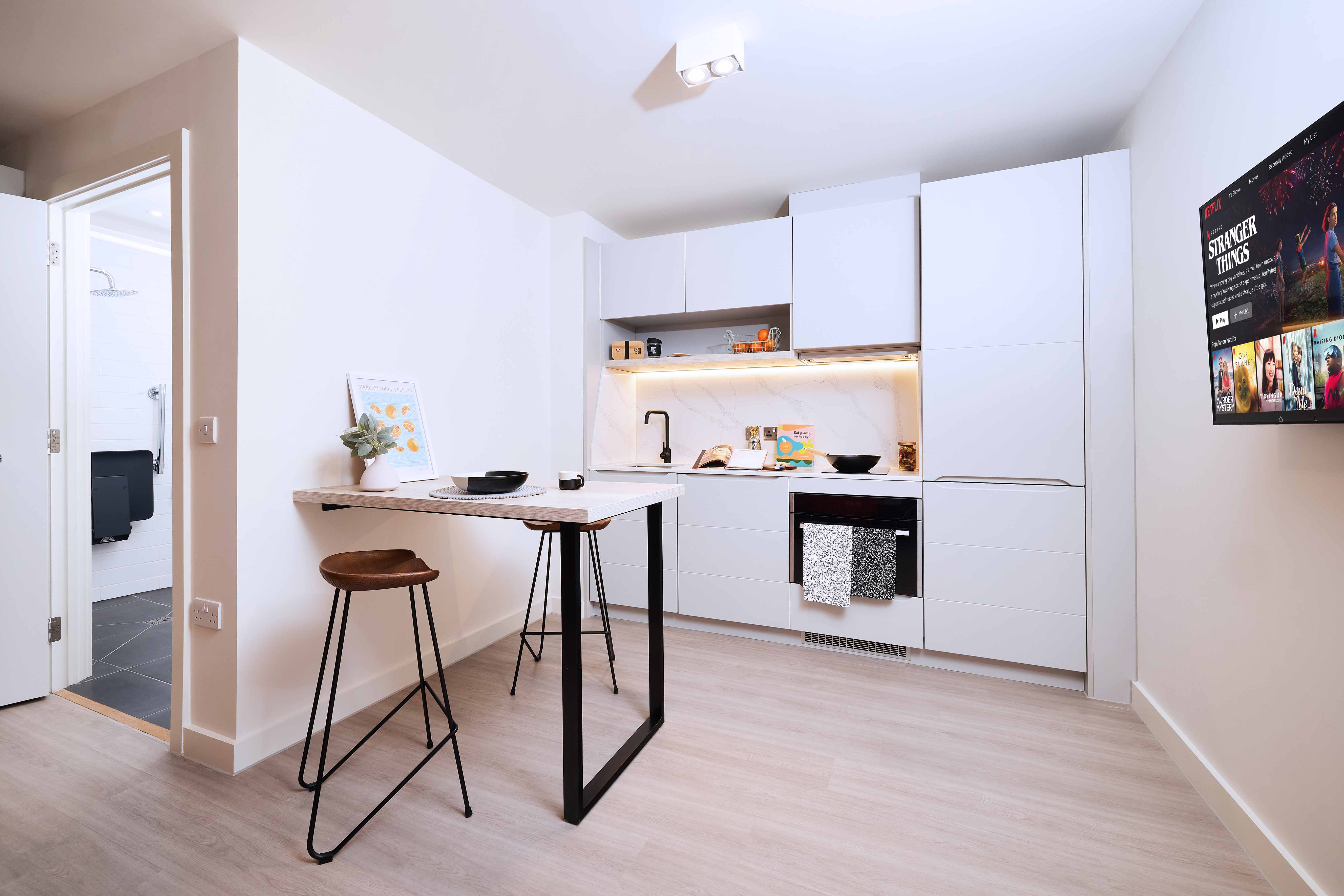 Modern kitchen with white cabinets and marble backsplash. A small island with two wooden stools is in the center. A wall-mounted TV is on the right. The open door reveals a bathroom. The space is bright with light wood flooring.