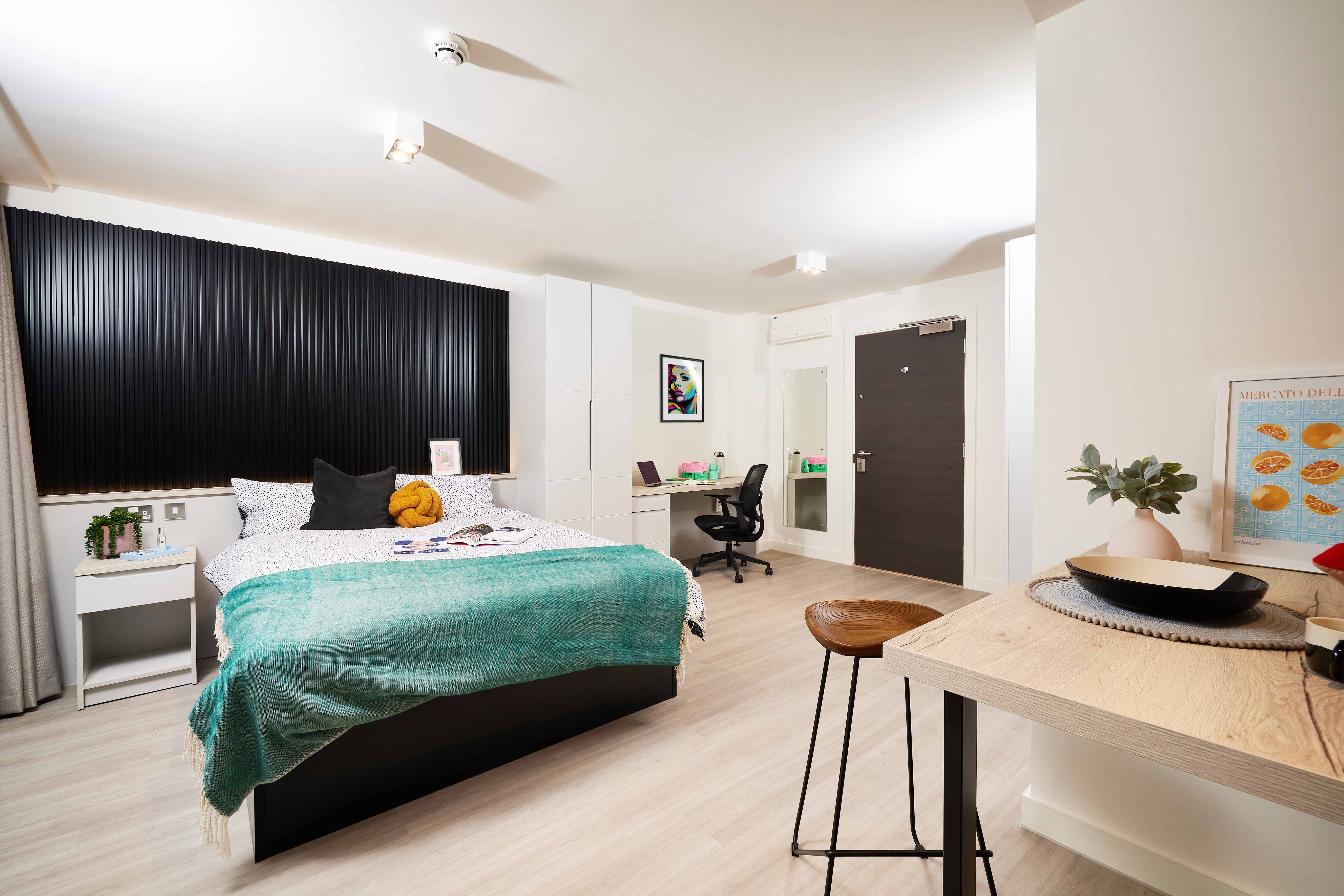 Modern bedroom with a double bed featuring a green blanket and decorative pillows. A black accent wall with a bookshelf above the bed. A study desk with a chair and a mirror, and a high table with stools are also visible. Minimalist decor throughout.