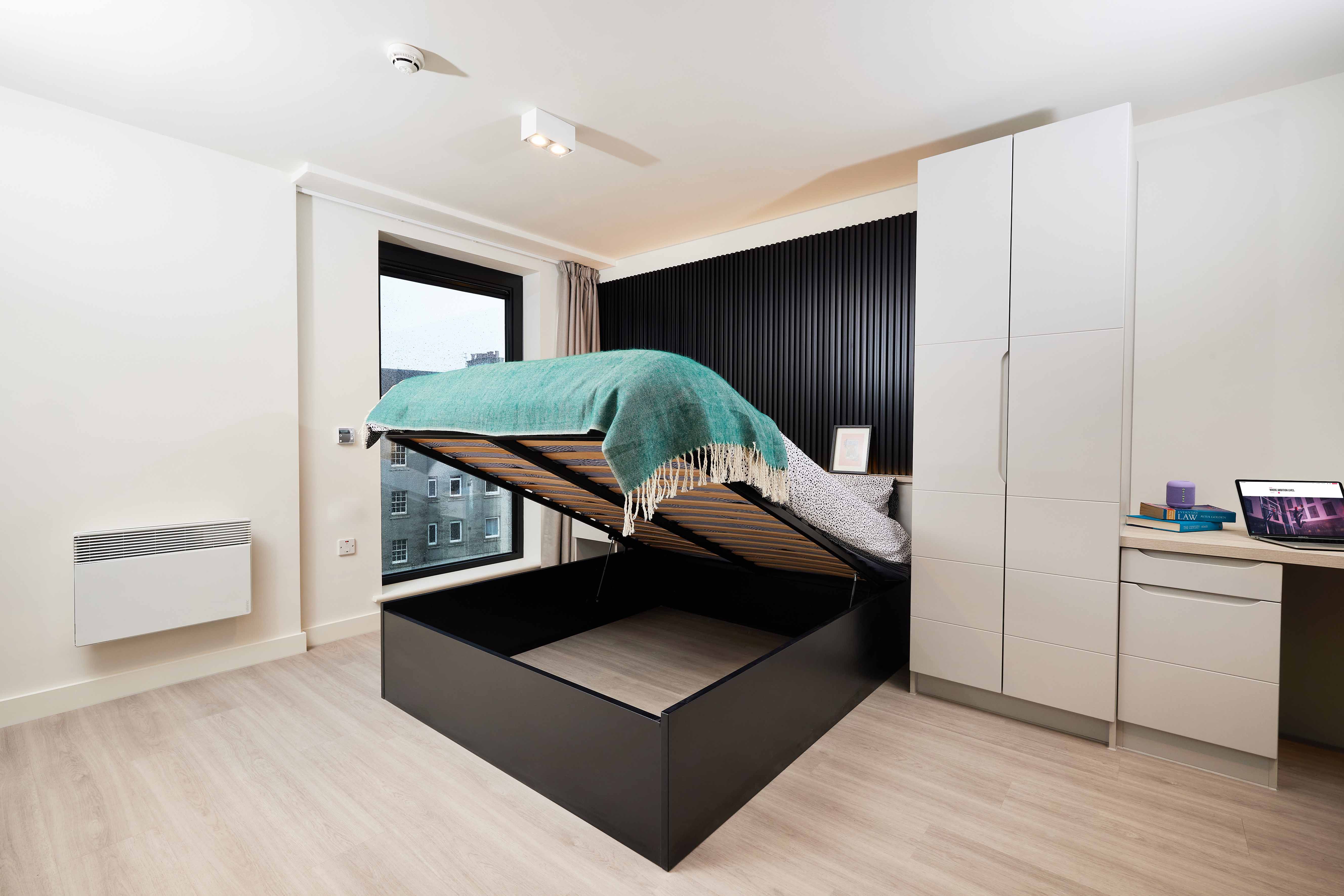 A modern bedroom with an open ottoman bed revealing storage underneath. The room has a large window, a white wardrobe, and a desk with a laptop. Natural light fills the space, showcasing light wood flooring and white walls.