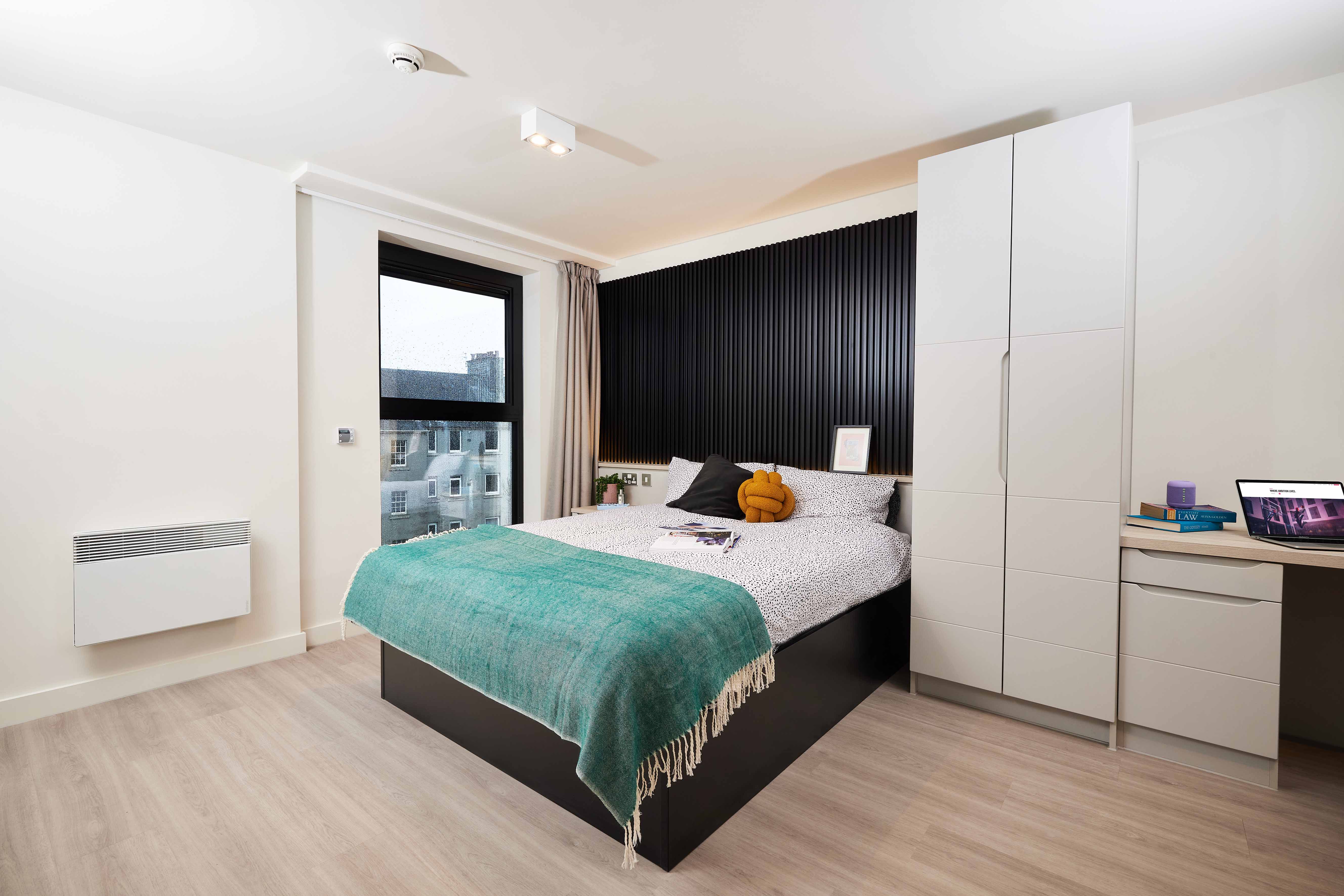 A modern bedroom with a large bed featuring a teal throw and mustard pillow. There's a black accent wall, a white wardrobe, a desk with a laptop and books, and a window with a view of buildings. The floor is light wood.