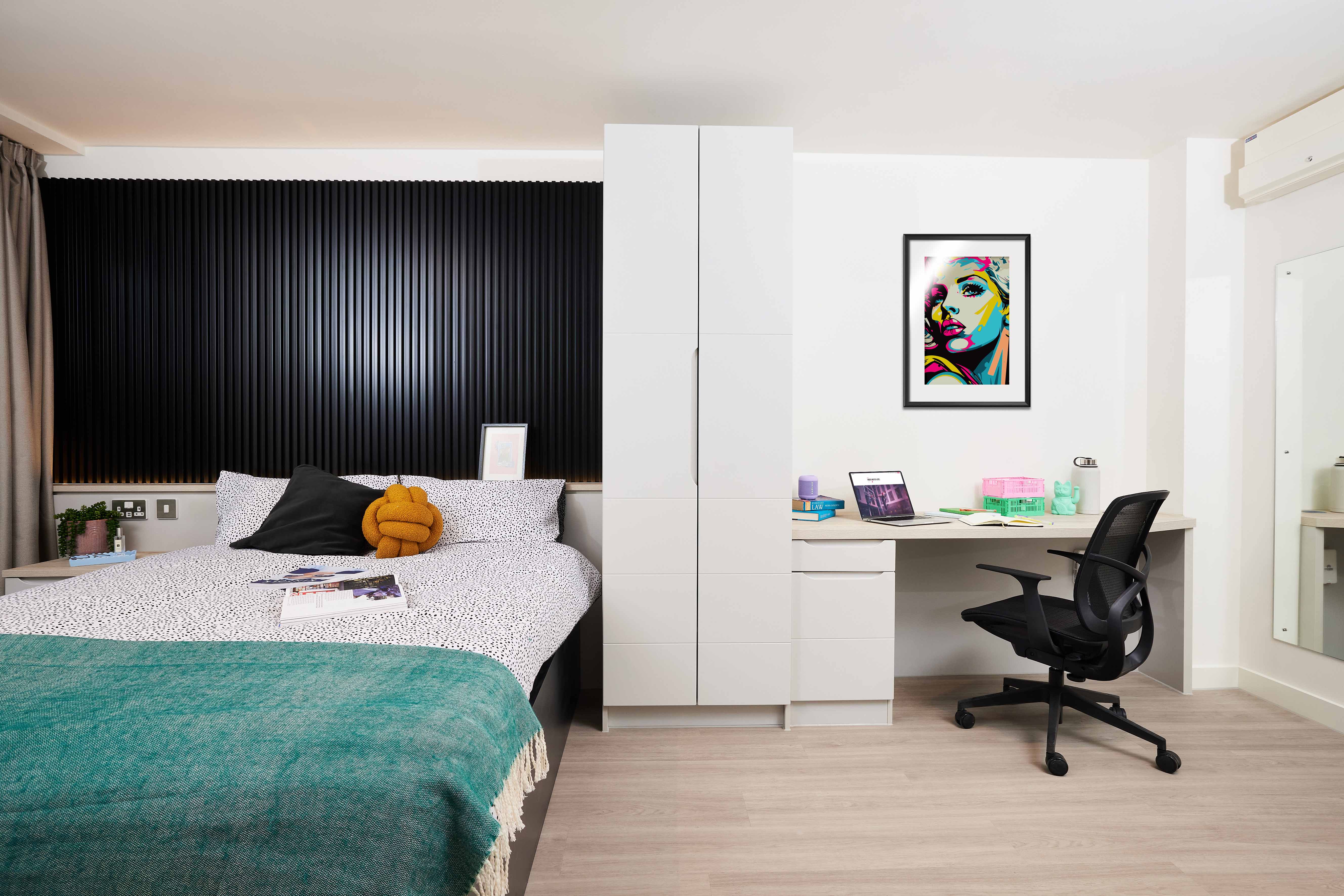 A modern bedroom with a bed, a turquoise throw, and a white cabinet. A desk with a laptop, books, and an office chair is on the right. A colorful portrait hangs on the wall. Light wood flooring and a black textured panel behind the bed complete the room.