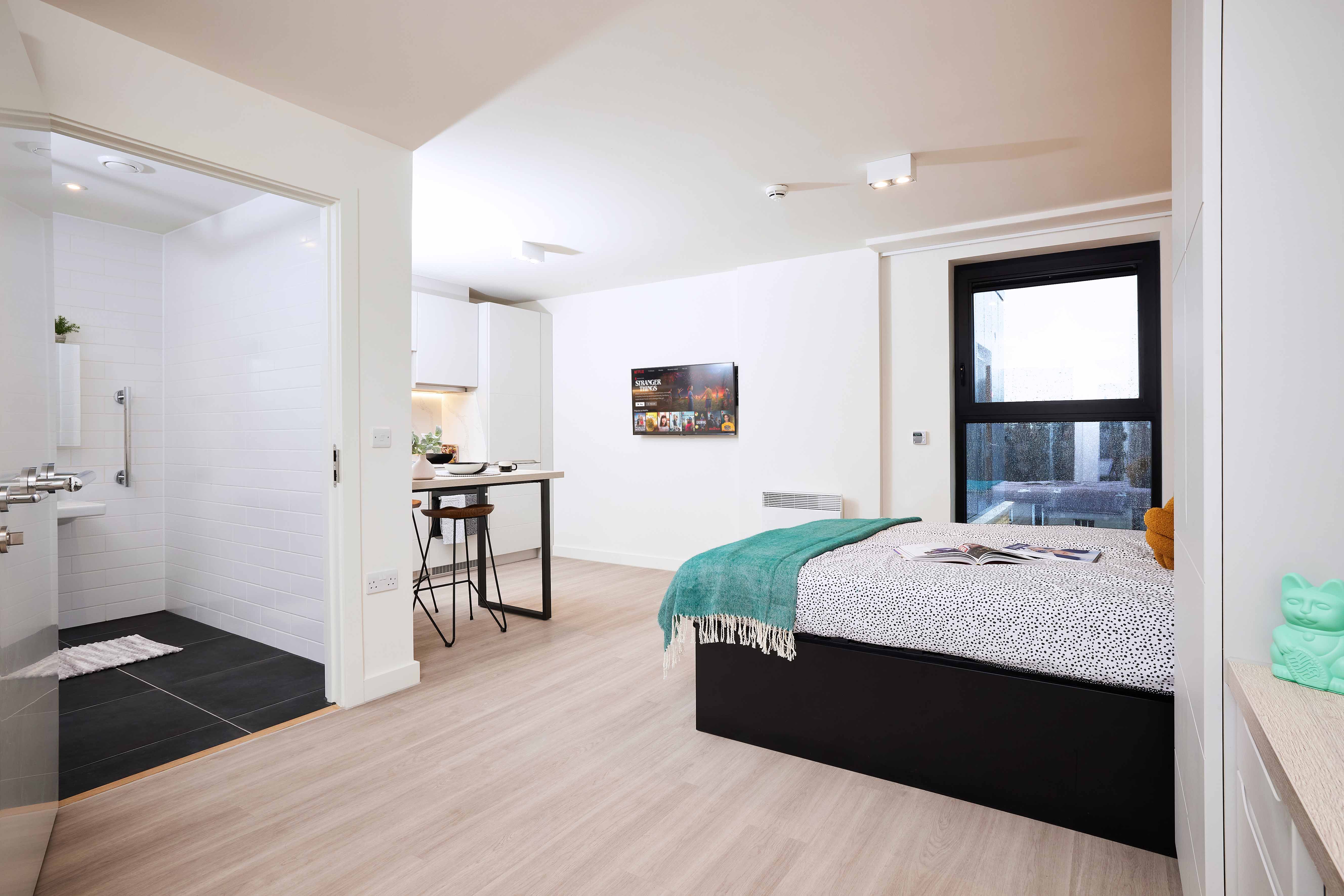 Modern studio apartment with light wood flooring, featuring a bed with a turquoise throw, a wall-mounted TV, a small kitchen area with a breakfast bar, and an adjacent bathroom with white tiles and a glass shower. A window provides natural light.