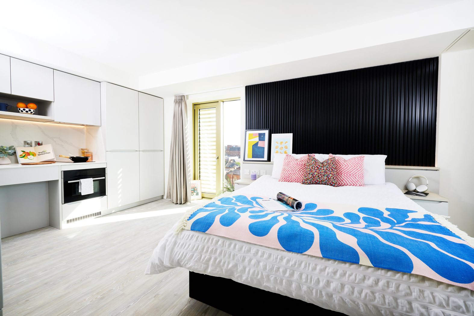A bright bedroom on Bruce Street features a white bed adorned with a colorful abstract blue blanket and pink cushions. The black-accented wall adds contrast. On the left, a kitchenette with white cabinets and an oven complements the space, while light curtains diffuse sunlight into the room.
