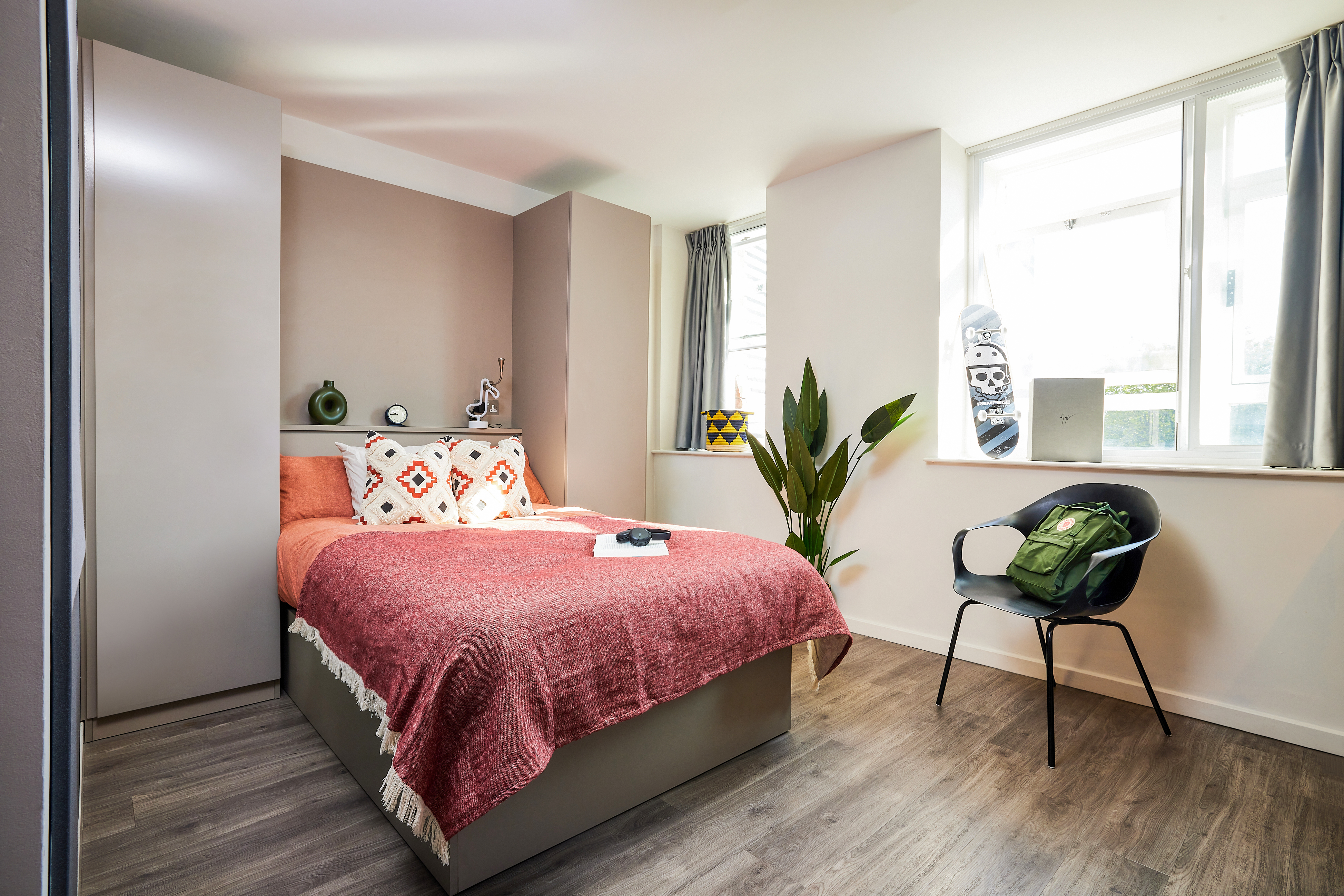 A cozy bedroom in a luxury student accommodation features a bed with a red blanket and decorative pillows. A plant is beside the bed, while a black chair holding a green backpack sits nearby. Light streams through two windows, and a skateboard leans against the wall.