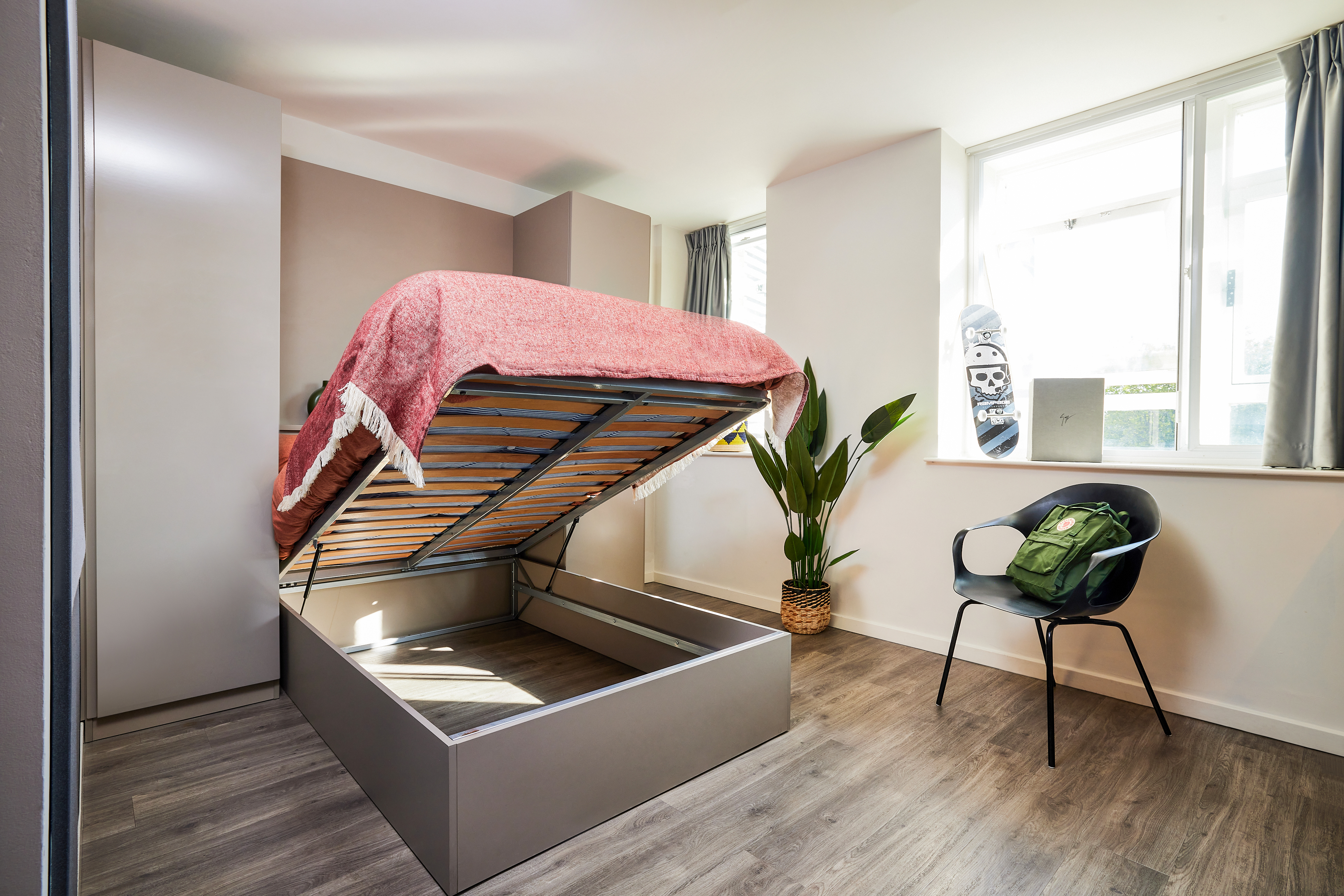 Experience luxury student accommodation with a room featuring a lifted platform storage bed adorned with a red blanket and pillows. A skateboard rests by the window, accompanied by a potted plant. A black chair holds a green backpack on the wooden floor, illuminated by natural light through the window.