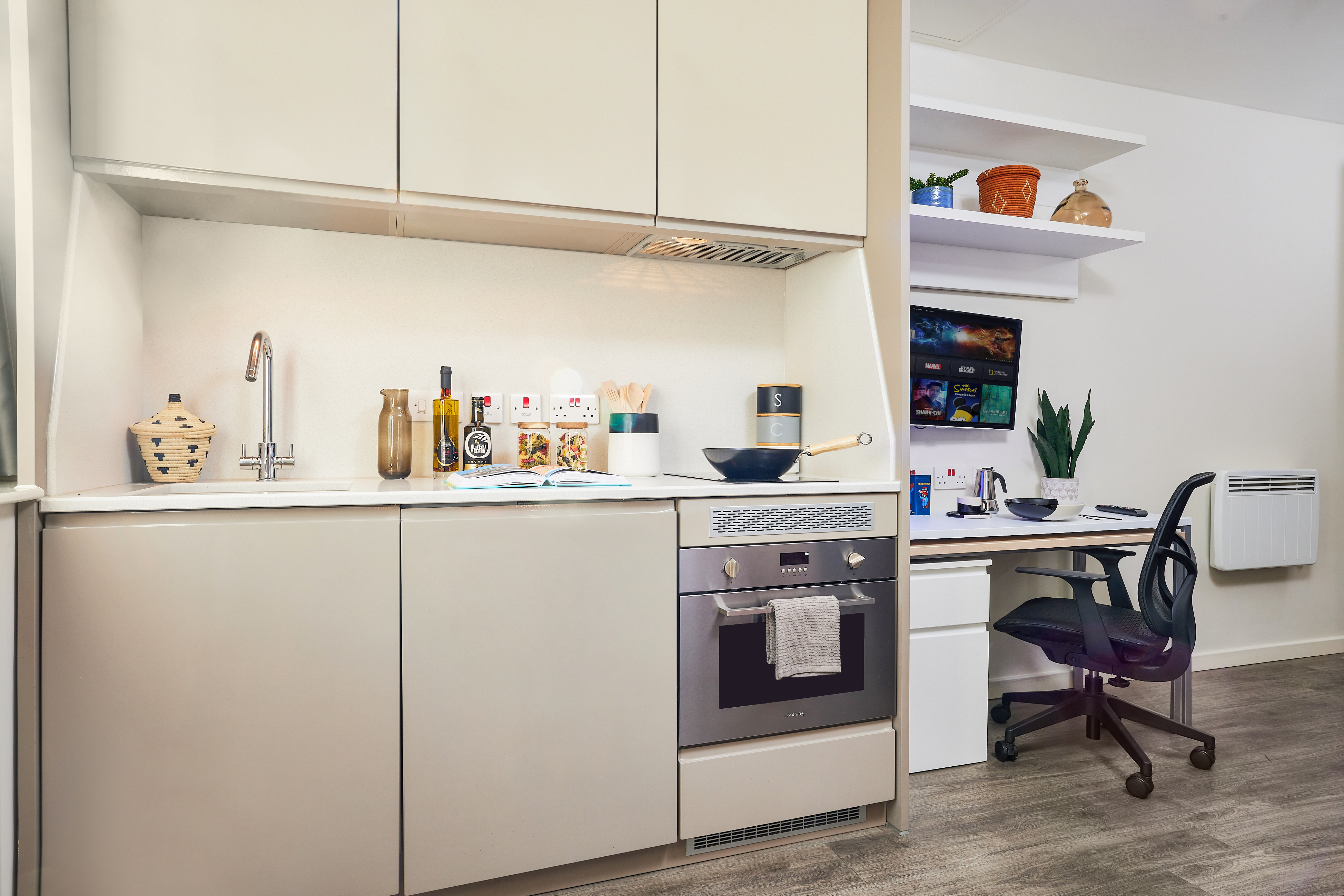 This luxury student accommodation features a modern studio apartment with a compact kitchen, including a sink, oven, and various cooking items. Adjacent is a workspace complete with a desk, chair, laptop, and wall shelves adorned with decorative items and a potted plant.