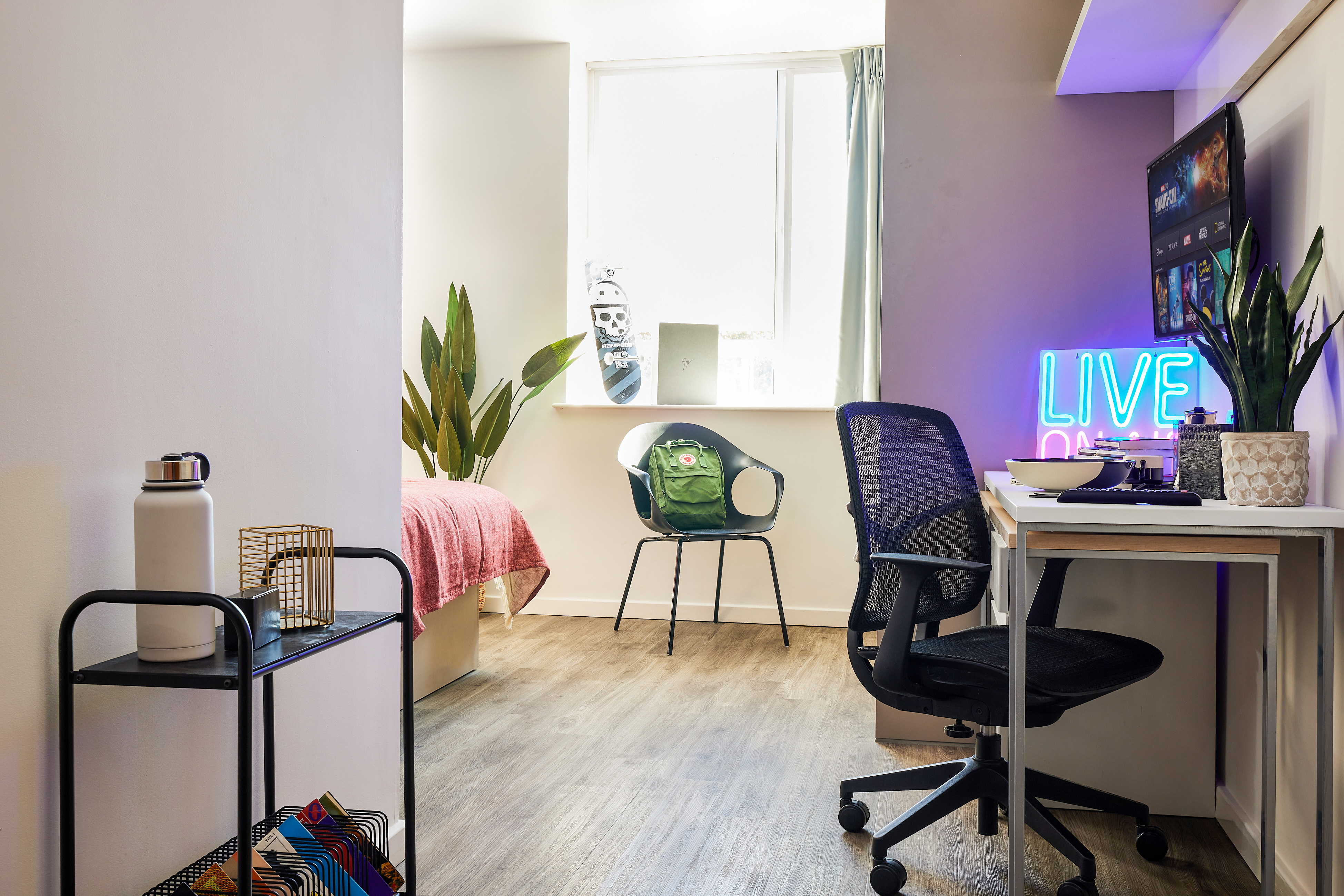 This luxury student accommodation features a modern room with a desk and computer. A neon "LIVE" sign adorns the wall. The chair faces a window by the bed with pink bedding, while a potted plant and a chair with a green bag complete the decor, complemented by wood flooring throughout.