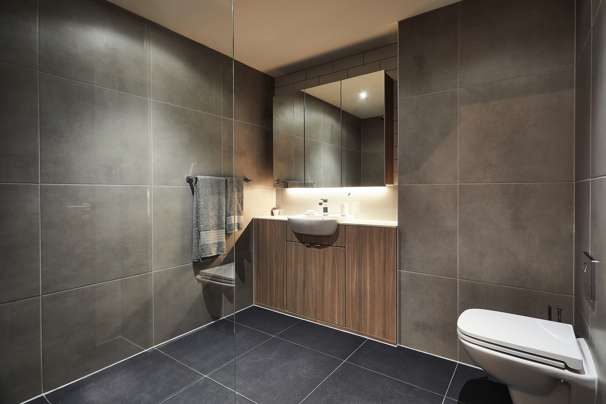 Modern bathroom on Lawrence Street with gray tiled walls and floor, featuring a wall-mounted toilet, a small sink with a mirror above, and a wooden cabinet. A towel hangs on the wall, and recessed lighting provides a warm ambiance.