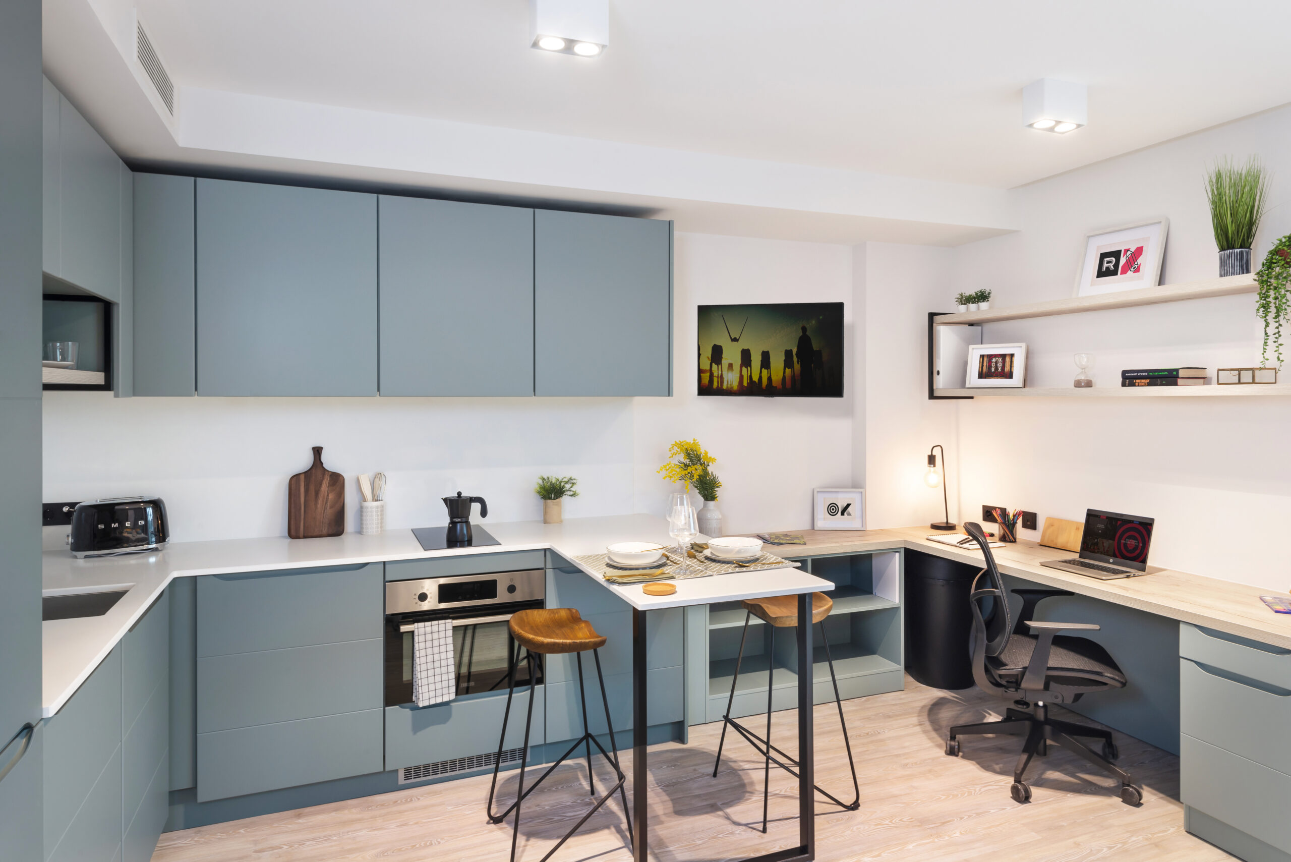 Located on Bruce Street, this modern kitchen and workspace features blue cabinets, an induction stove, and an oven on the left. On the right, a cozy nook offers a wooden table with stools and a computer workstation. Shelves are adorned with plants and books under bright ceiling lighting.